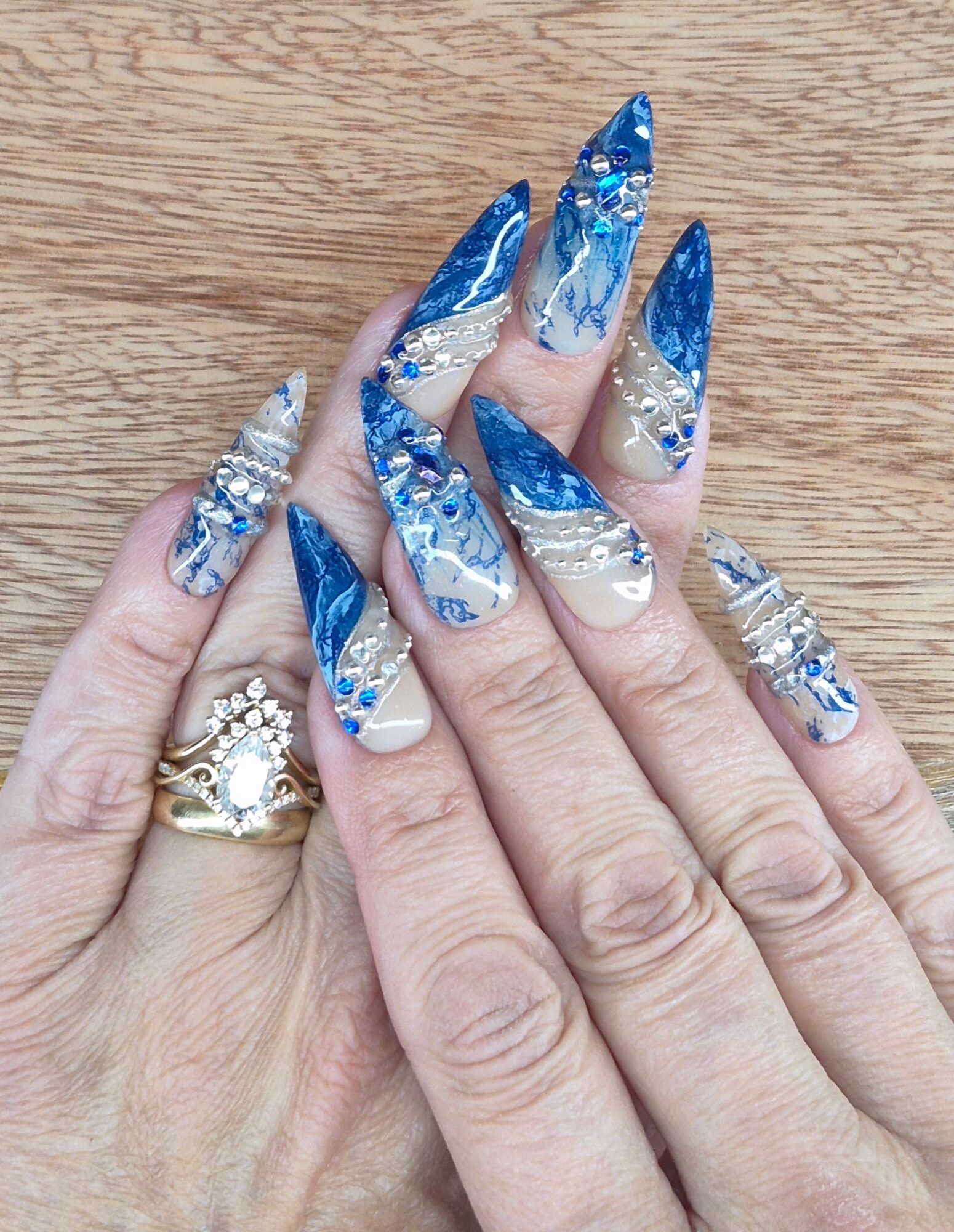 Hands with long, decorated nails featuring blue, white, and silver designs, and a gold ring with a large gemstone.