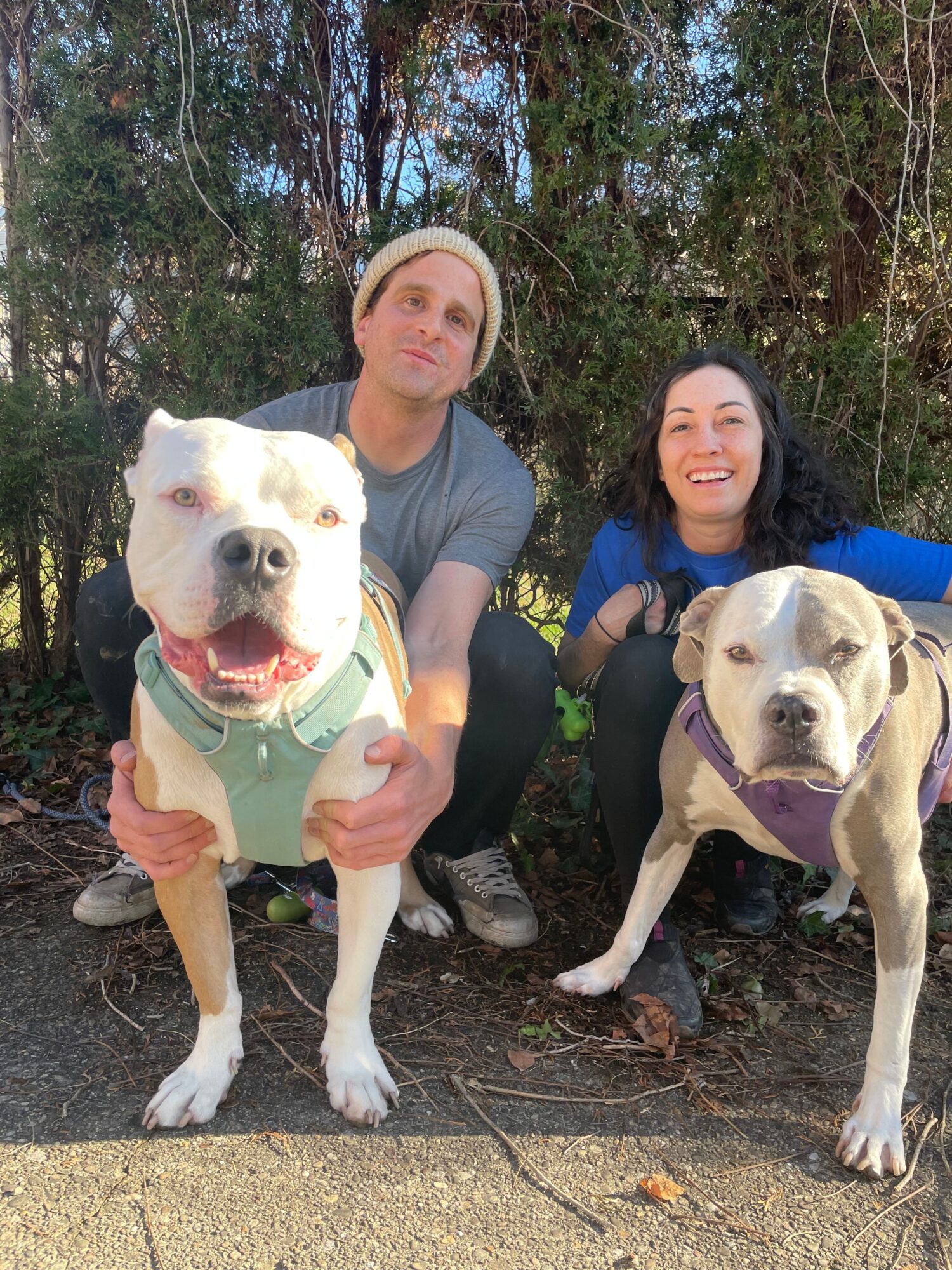 Two women and two dogs outdoors on a trail, with trees in the background. One woman is crouching, and the dogs are sitting nearby.