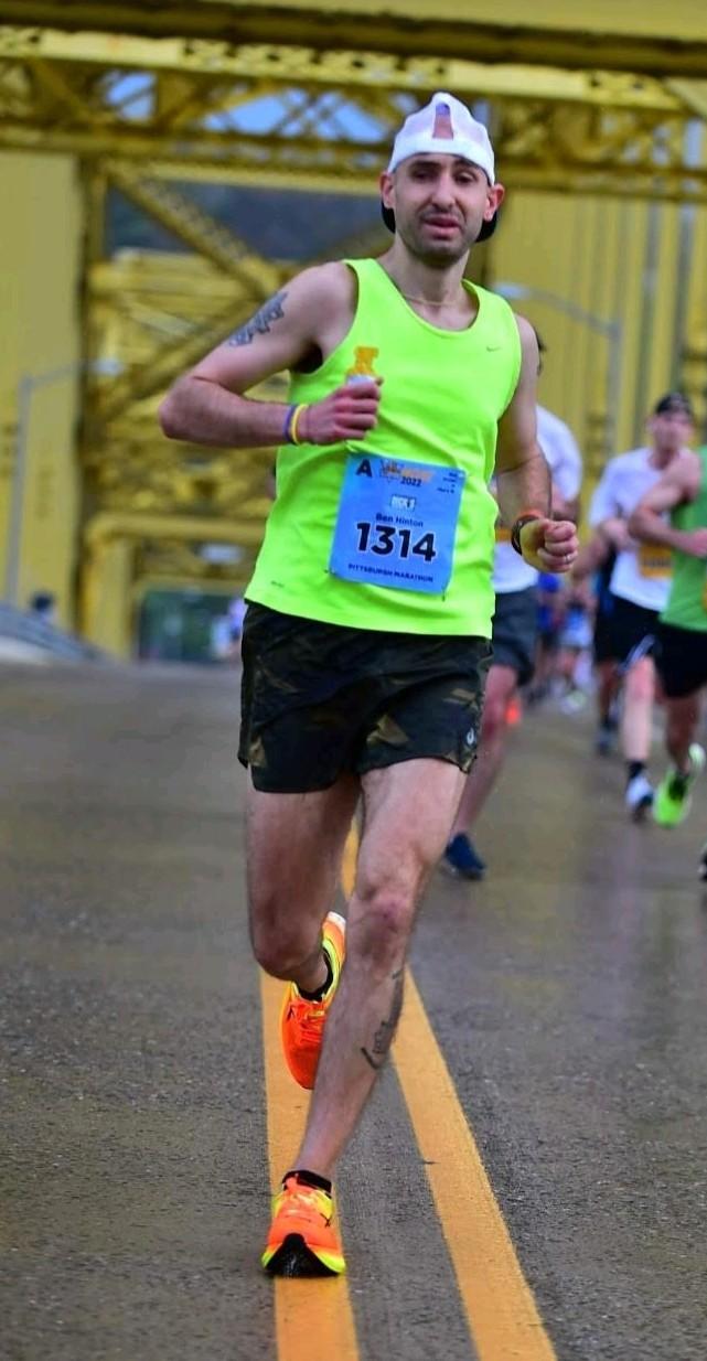 Male runner in neon green shirt and black shorts running on a bridge during a race, holding a water bottle, with other runners in background