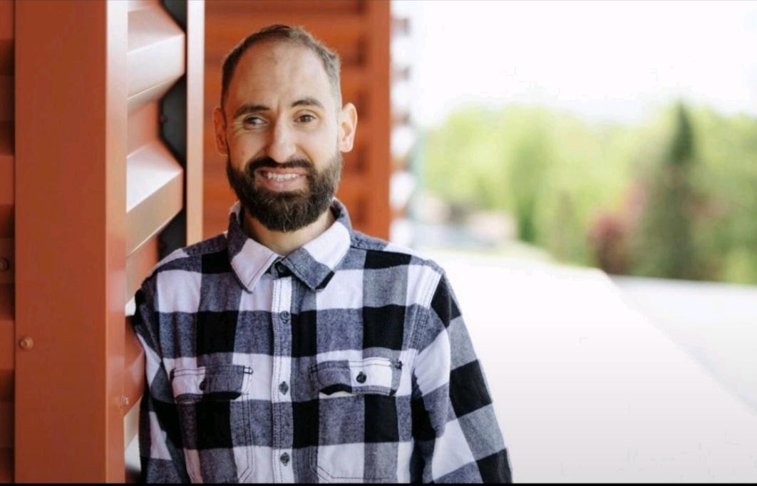 Man with a beard smiling outdoors, wearing a checkered shirt, standing near a wooden structure, with blurred greenery in background.