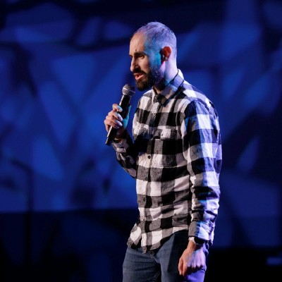 Man with a beard and short hair speaking into a microphone on stage, wearing a checkered shirt, with a blue background.