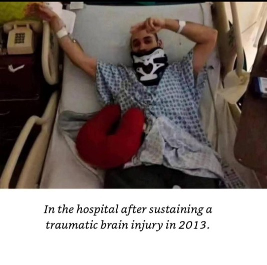 Person lying in hospital bed with arm raised, wearing a mask, with a red pillow, hospital equipment nearby, smiling.