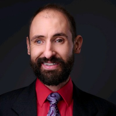 Man with a beard, wearing a suit, red shirt, and patterned tie, smiling against a dark background.
