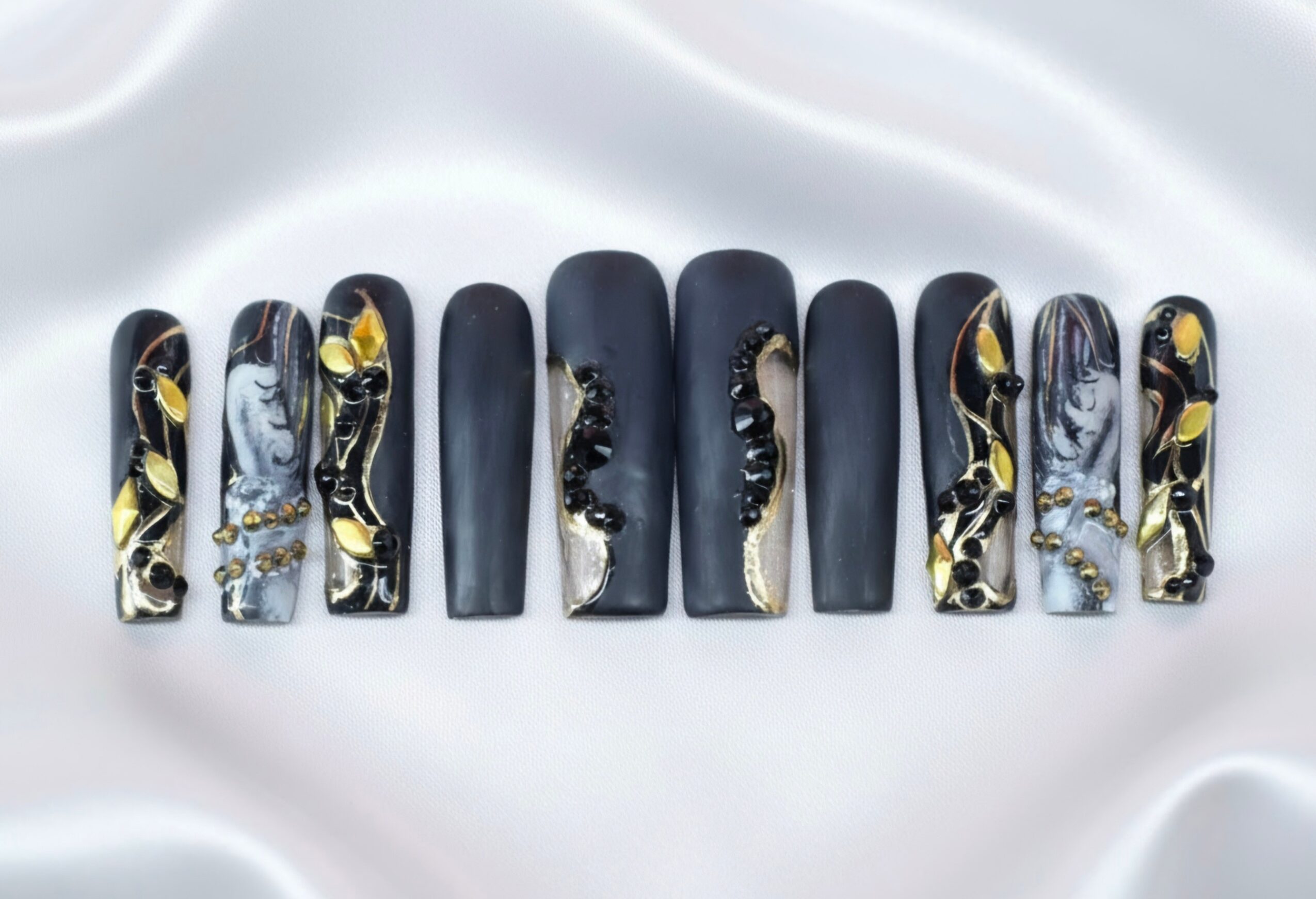 Set of ten artificial nails with black, gold, and silver designs on a white satin background.