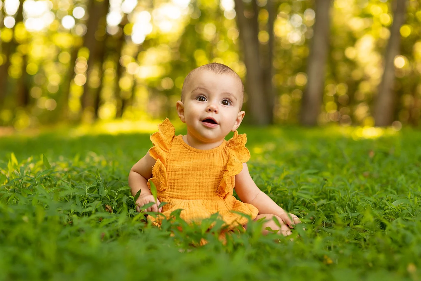 Baby girl sitting on grass outdoors with trees in background, wearing a yellow dress, looking at camera, smiling.