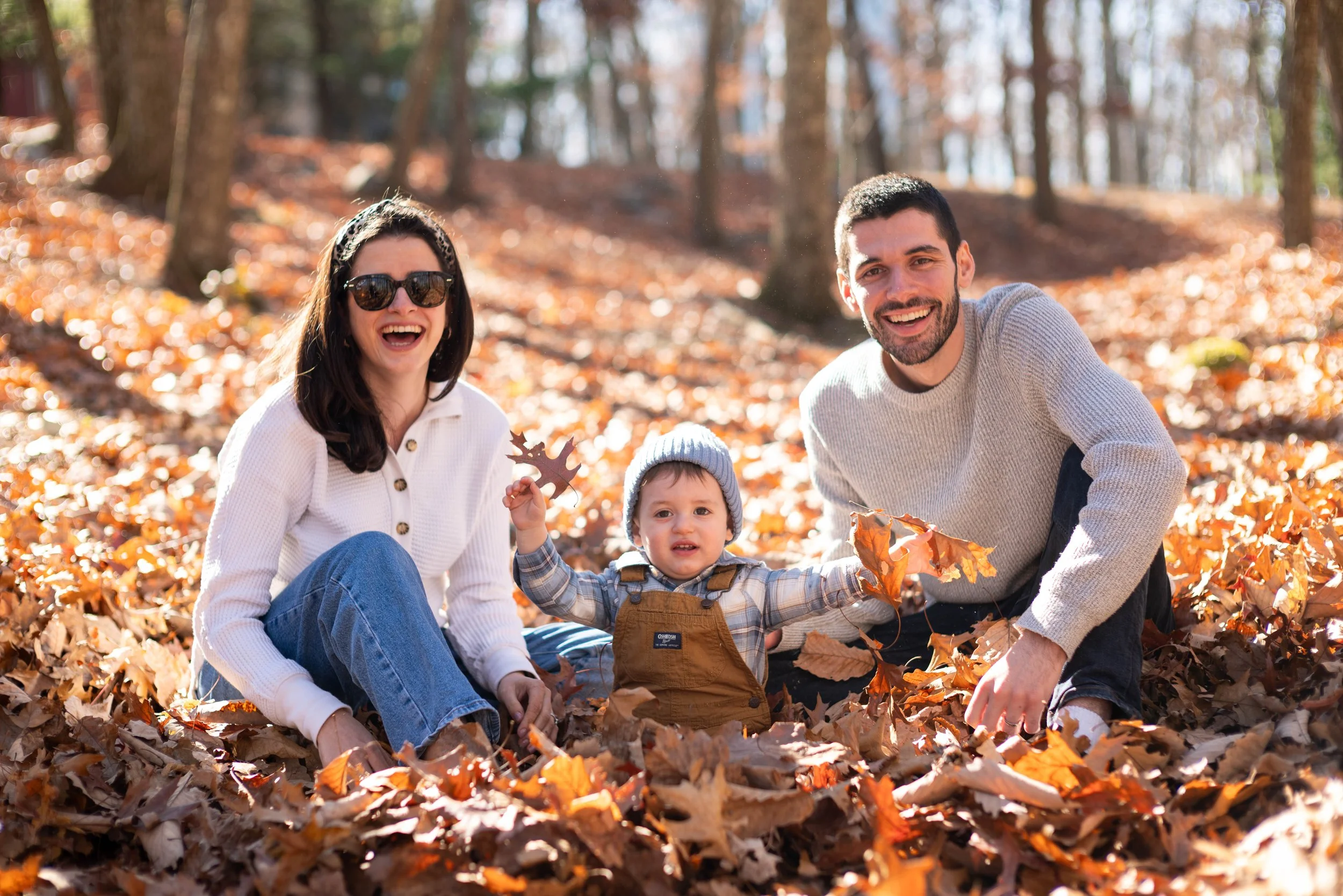 Family of three sitting outdoors on fallen leaves in a forest, smiling and playing, with trees in the background.