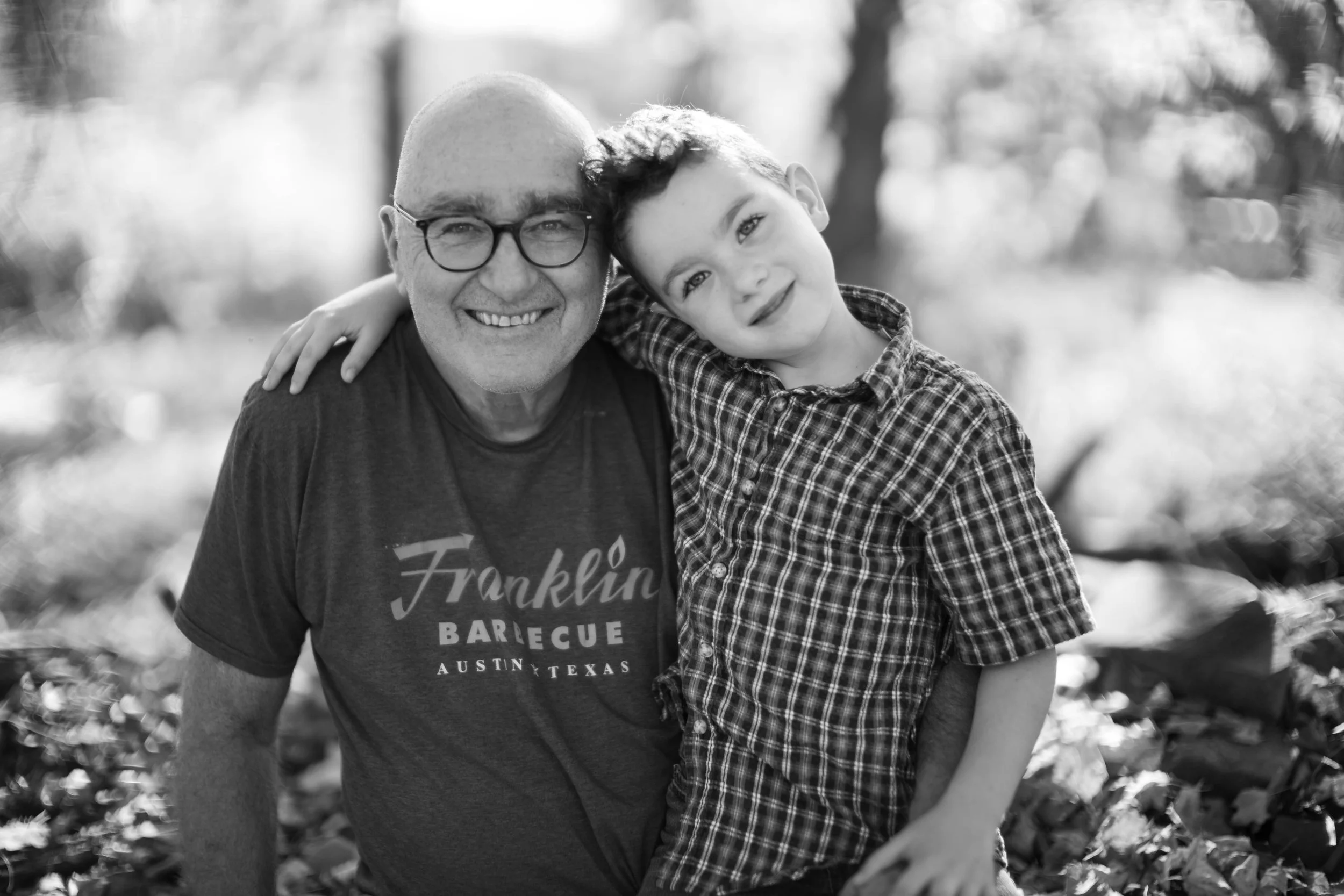 A man and a boy smile and hug outdoors, with trees and sunlight in the background.