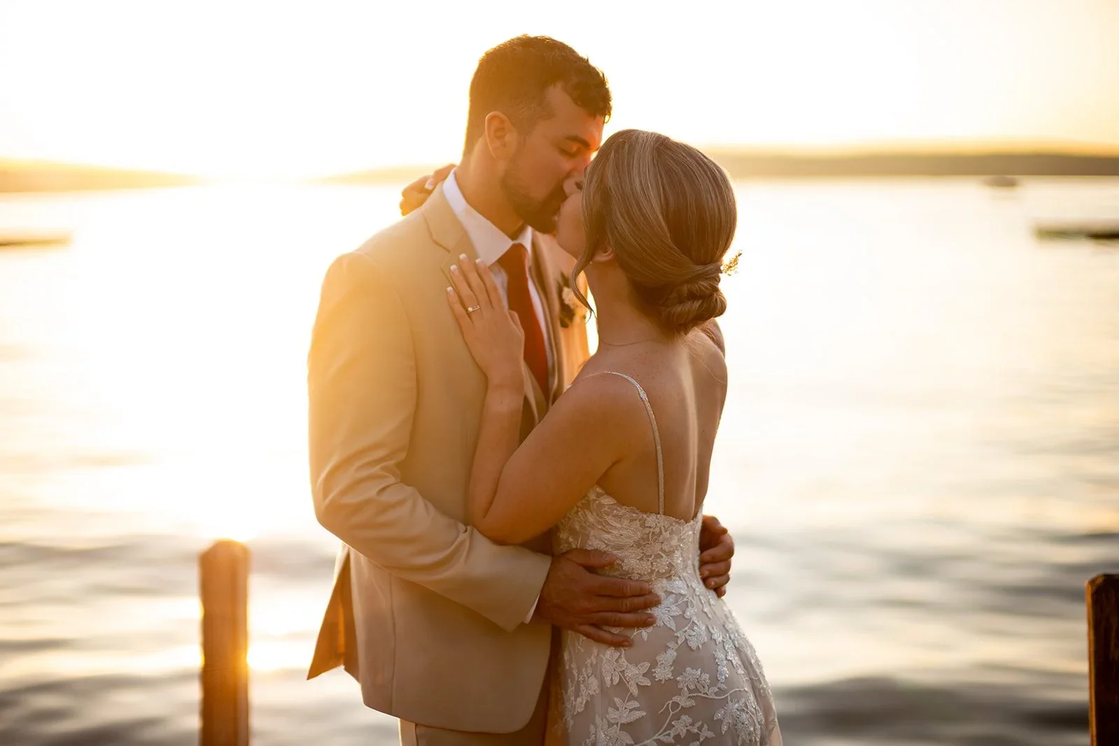 A couple in wedding attire embraces by a body of water during sunset, with the man in a suit and the woman in a lace dress.