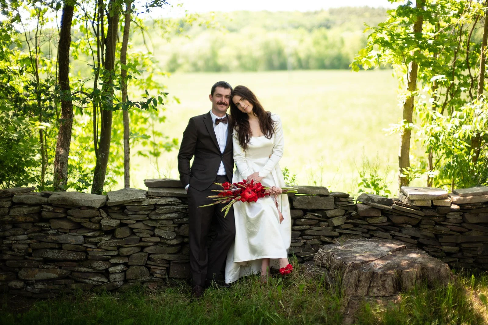 A couple dressed in formal attire standing outdoors near a stone wall, with trees and a field in the background.