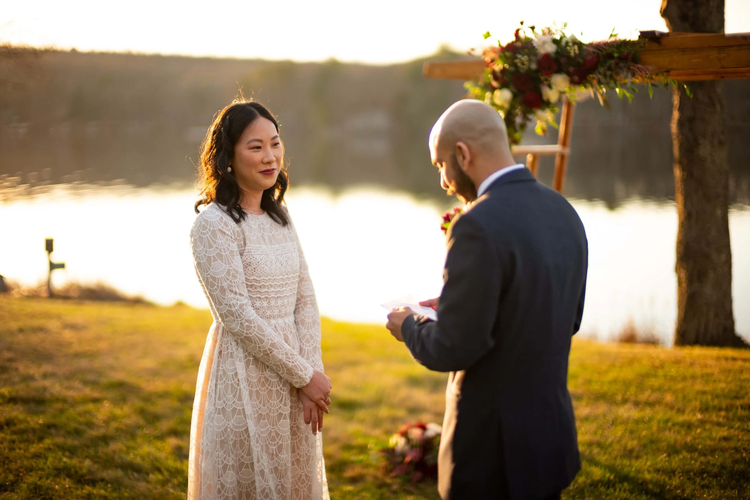 A woman in a white dress stands outdoors near a lake, facing a man in a suit during sunset.