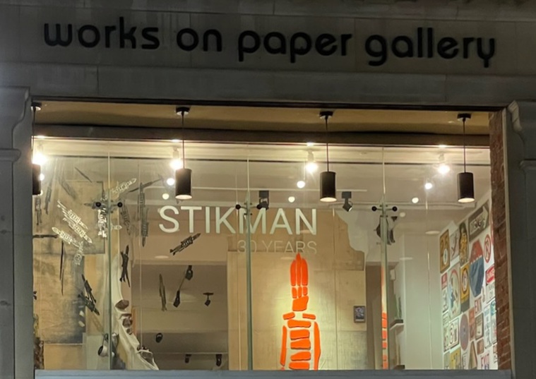 Store window displays artwork and text, with lights illuminating the interior, and a sign reading 'works on paper gallery' above.