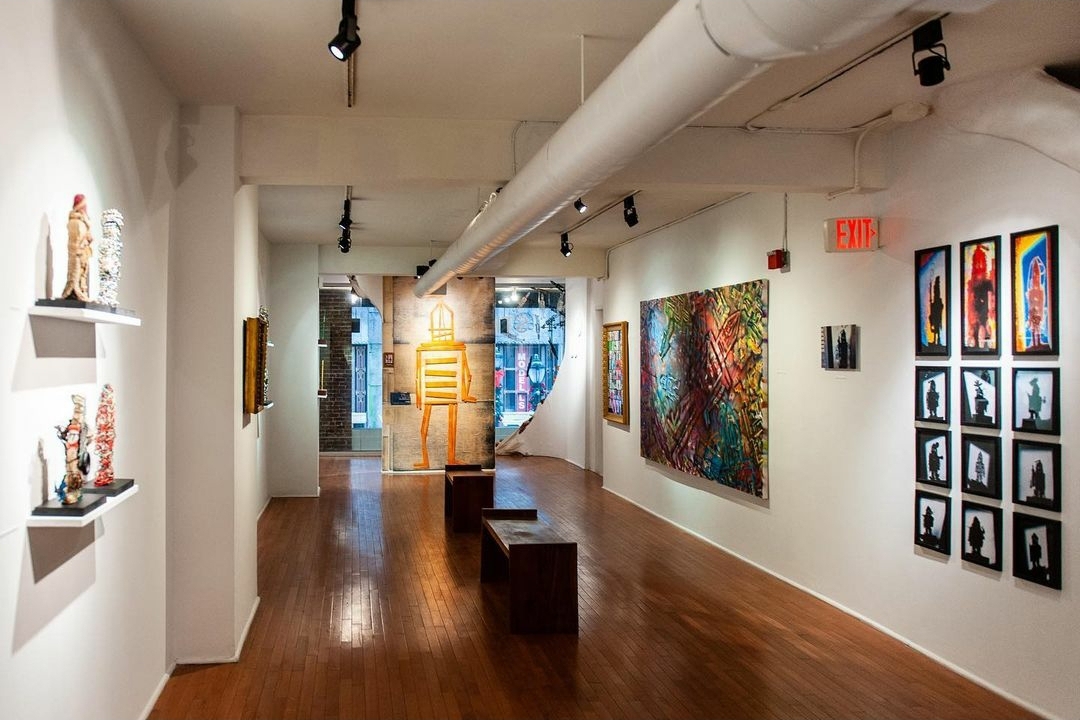 Gallery interior with colorful artwork on white walls, wooden floor, and ceiling track lighting, viewed from the entrance.