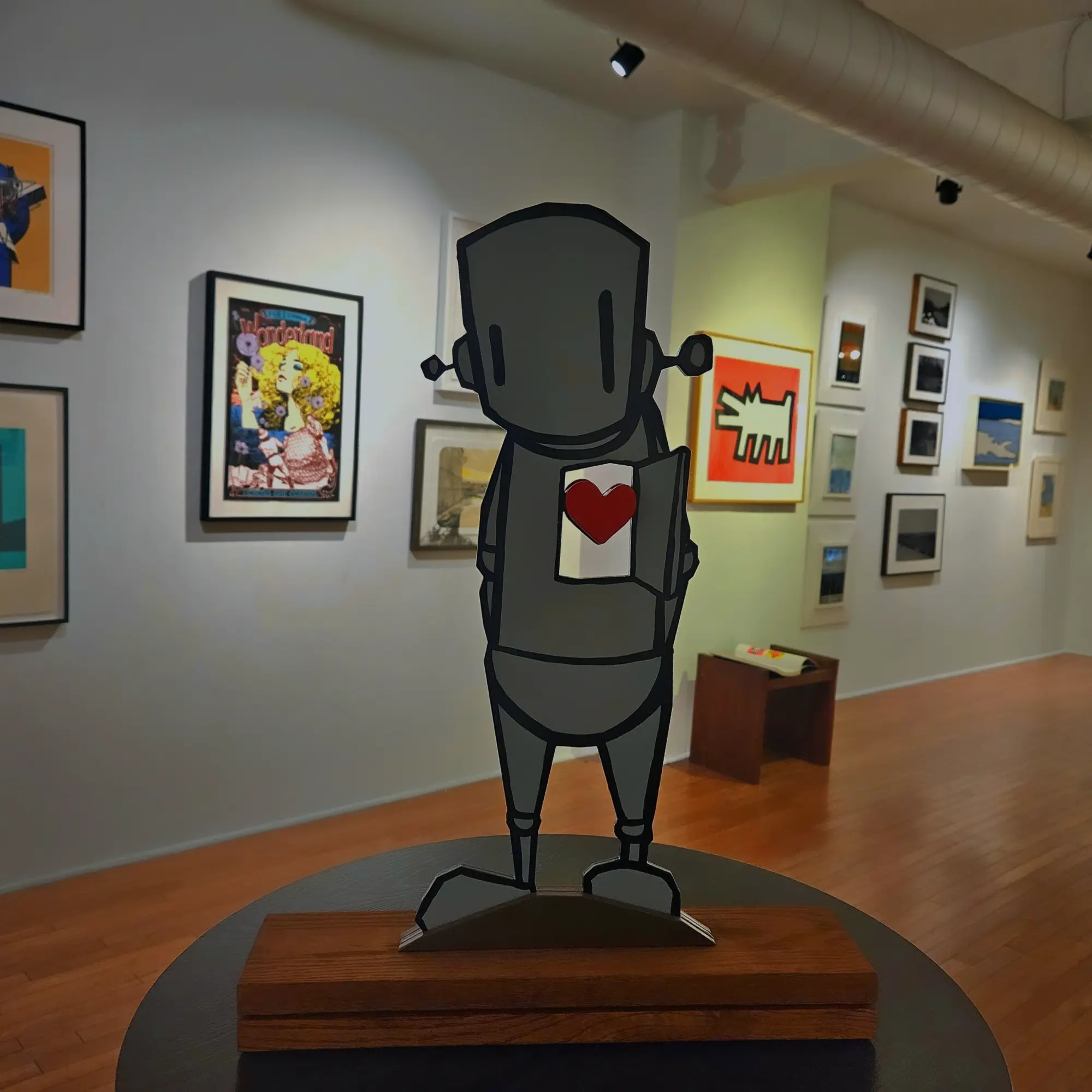 A robot sculpture with a heart on its chest stands on a wooden base in an art gallery.