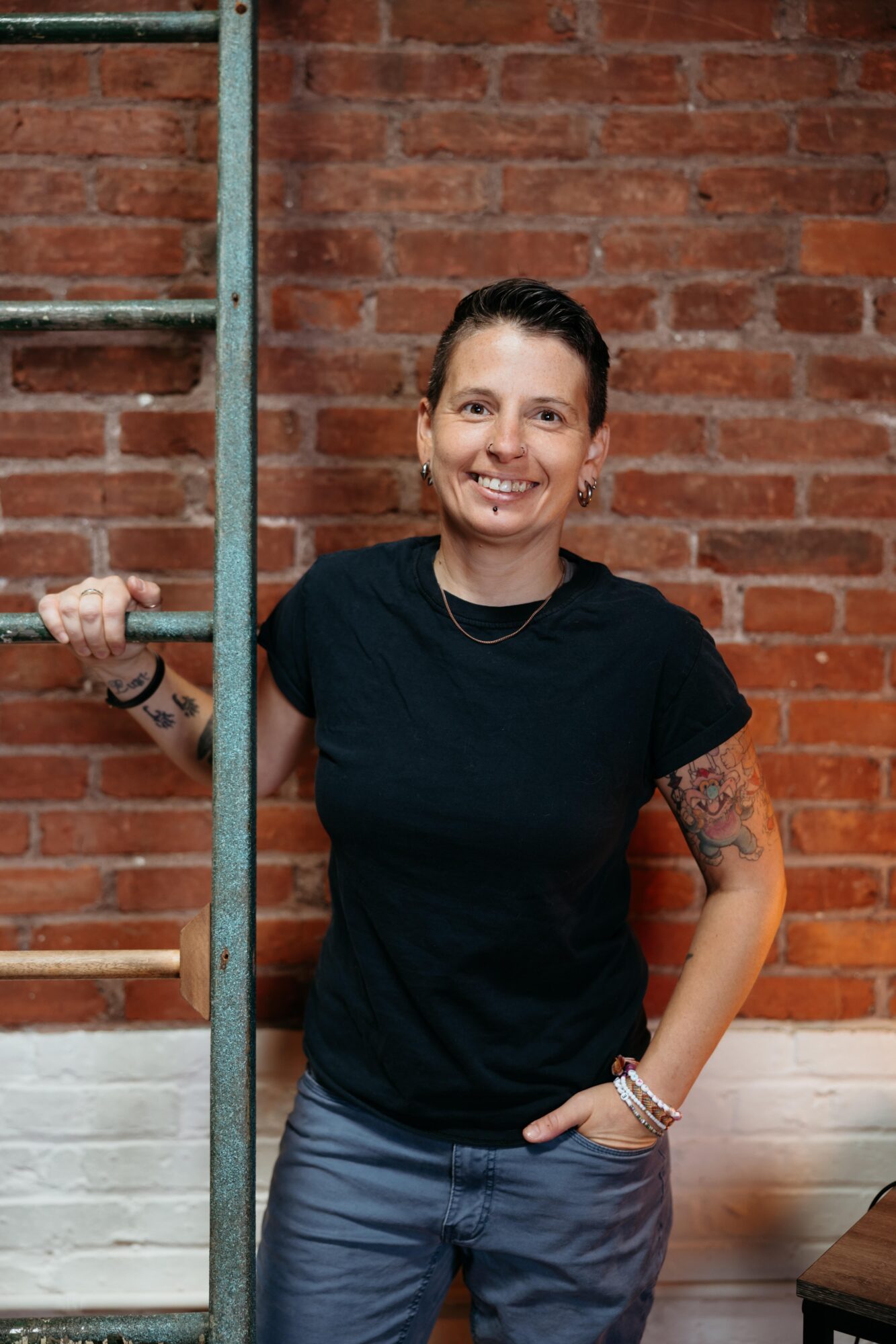 Smiling woman with short dark hair, tattoos on arms, wearing a black t-shirt and jeans, standing against a brick wall.