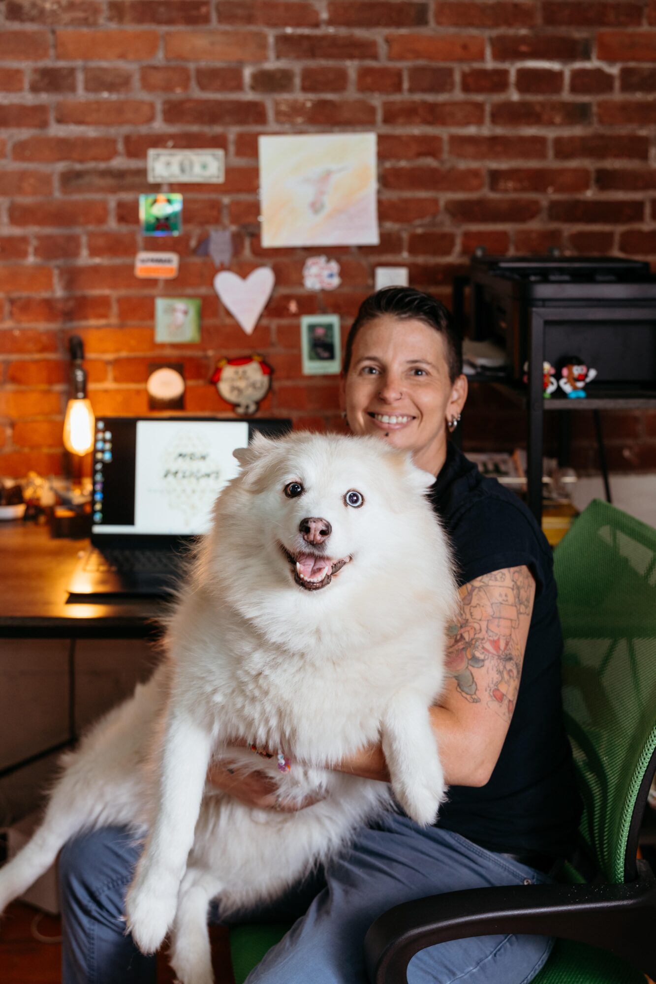 Person smiling holding a white fluffy dog indoors with a brick wall background and colorful drawings