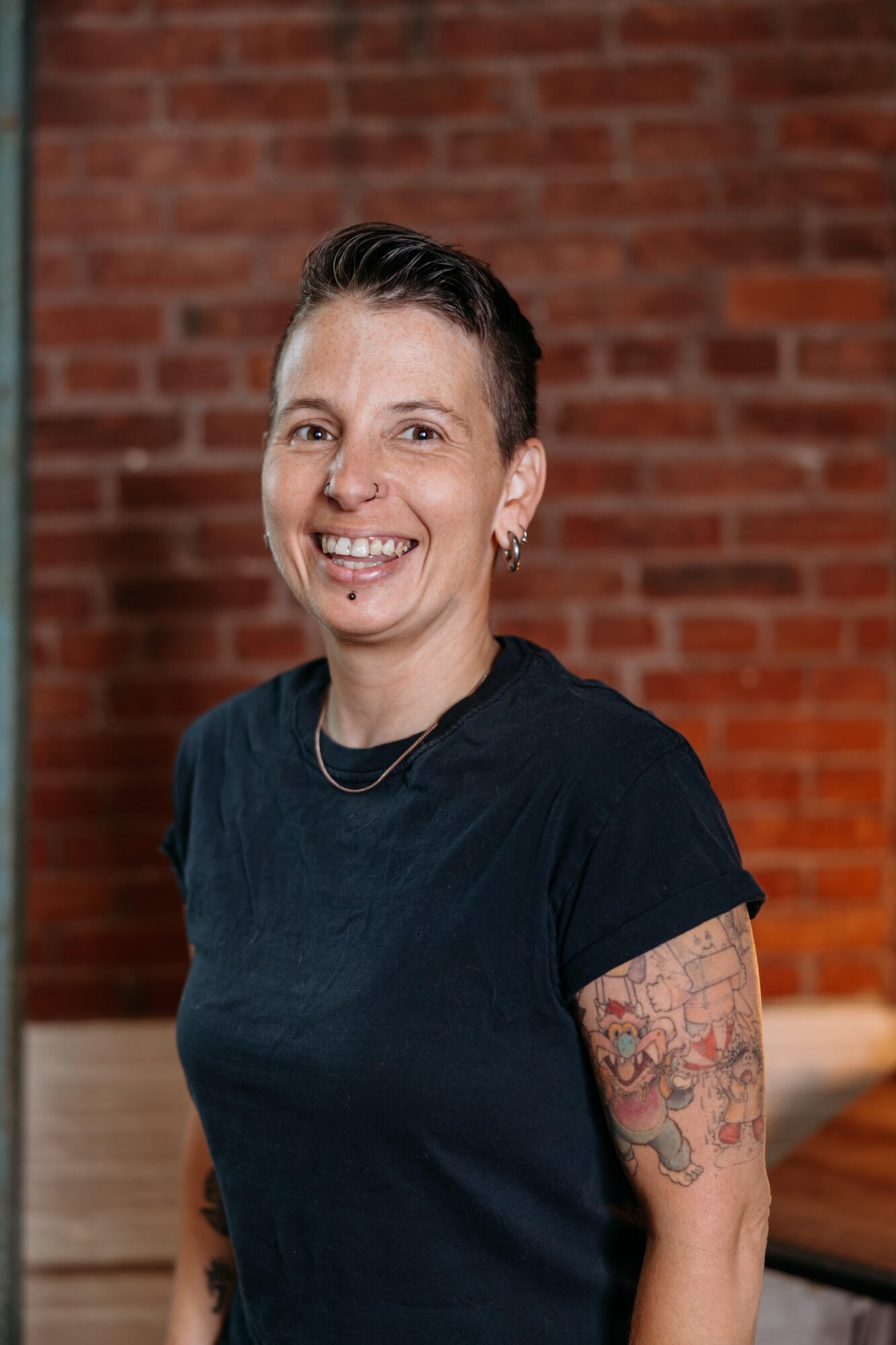 Smiling woman with short dark hair and tattoos on her left arm, wearing a black shirt, standing in front of a brick wall.