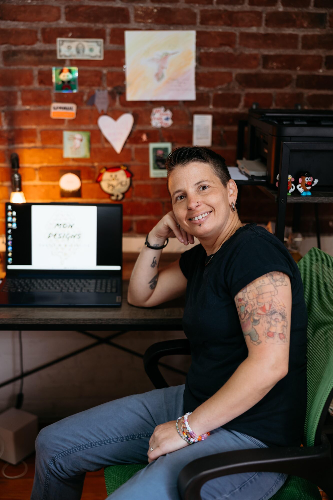Person with short hair and tattoos sitting at a desk with a laptop, smiling, against a brick wall decorated with photos and artwork.