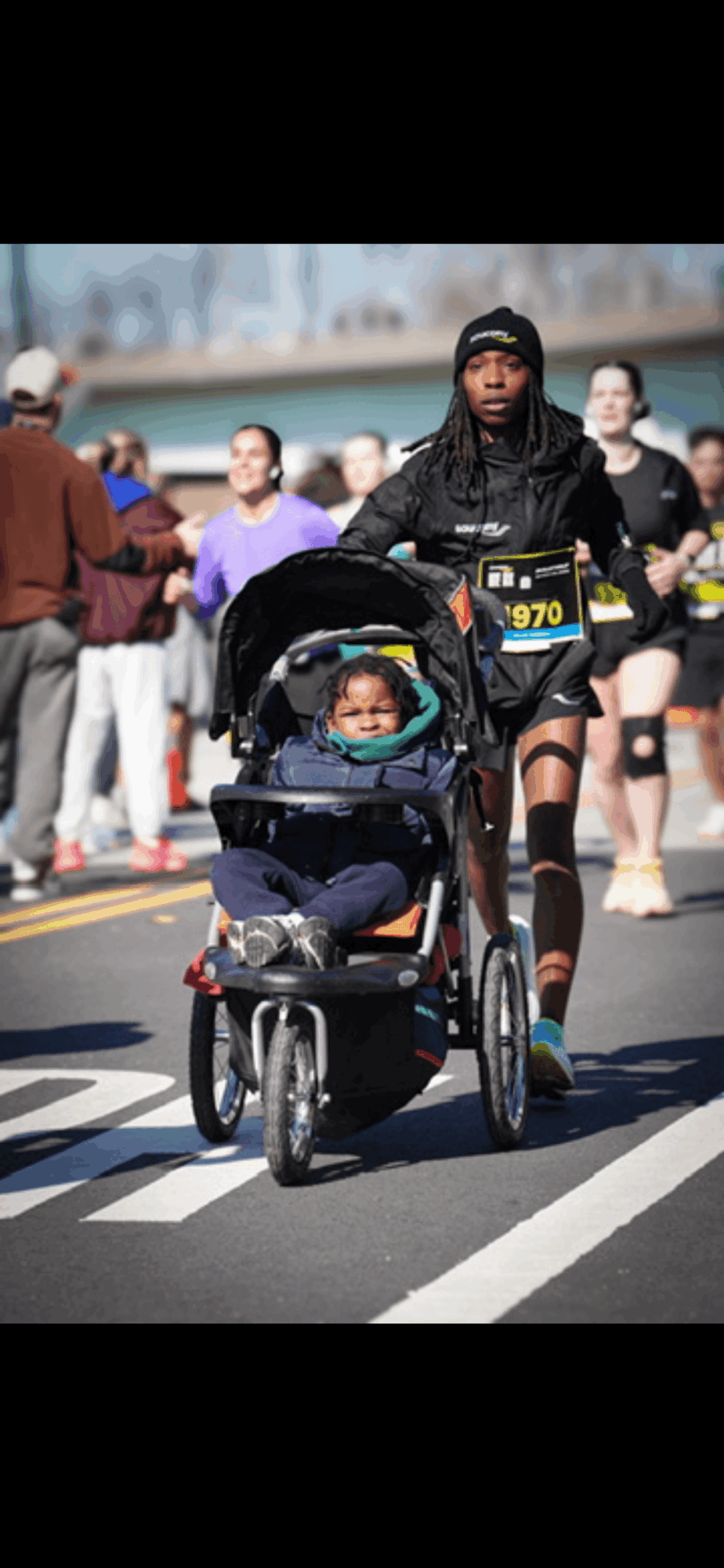 Person riding a bicycle with a child in a stroller, surrounded by runners and spectators on a street.