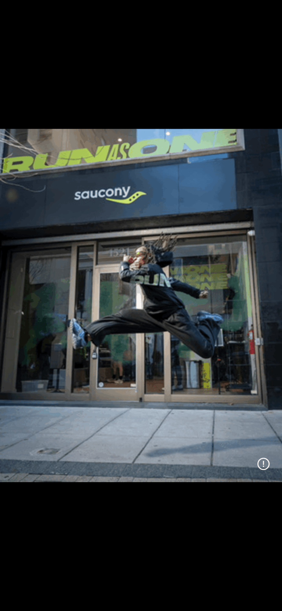 Person mid-air jumping in front of a store entrance with glass doors and a Saucony sign above.
