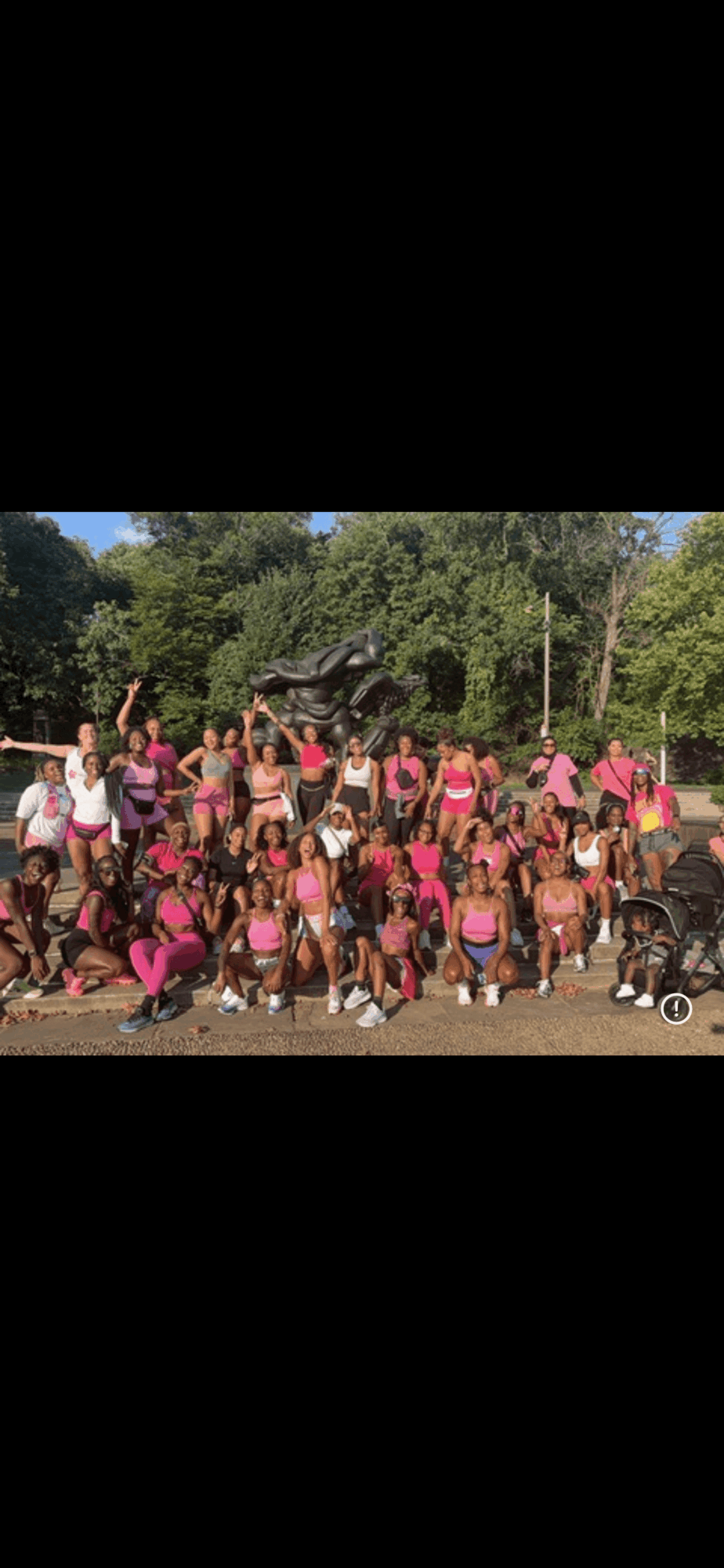 Group of people outdoors, some wearing pink, with trees and a statue in the background.