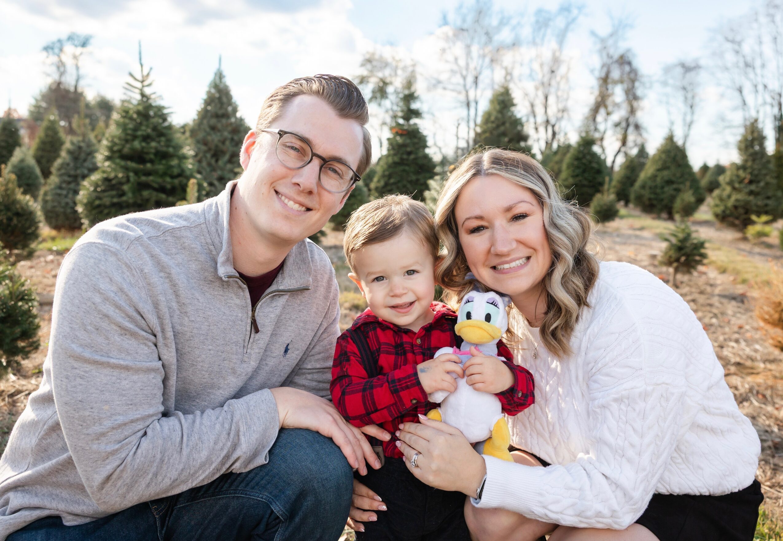 Family of three smiling outdoors, holding a plush toy, with trees in the background.