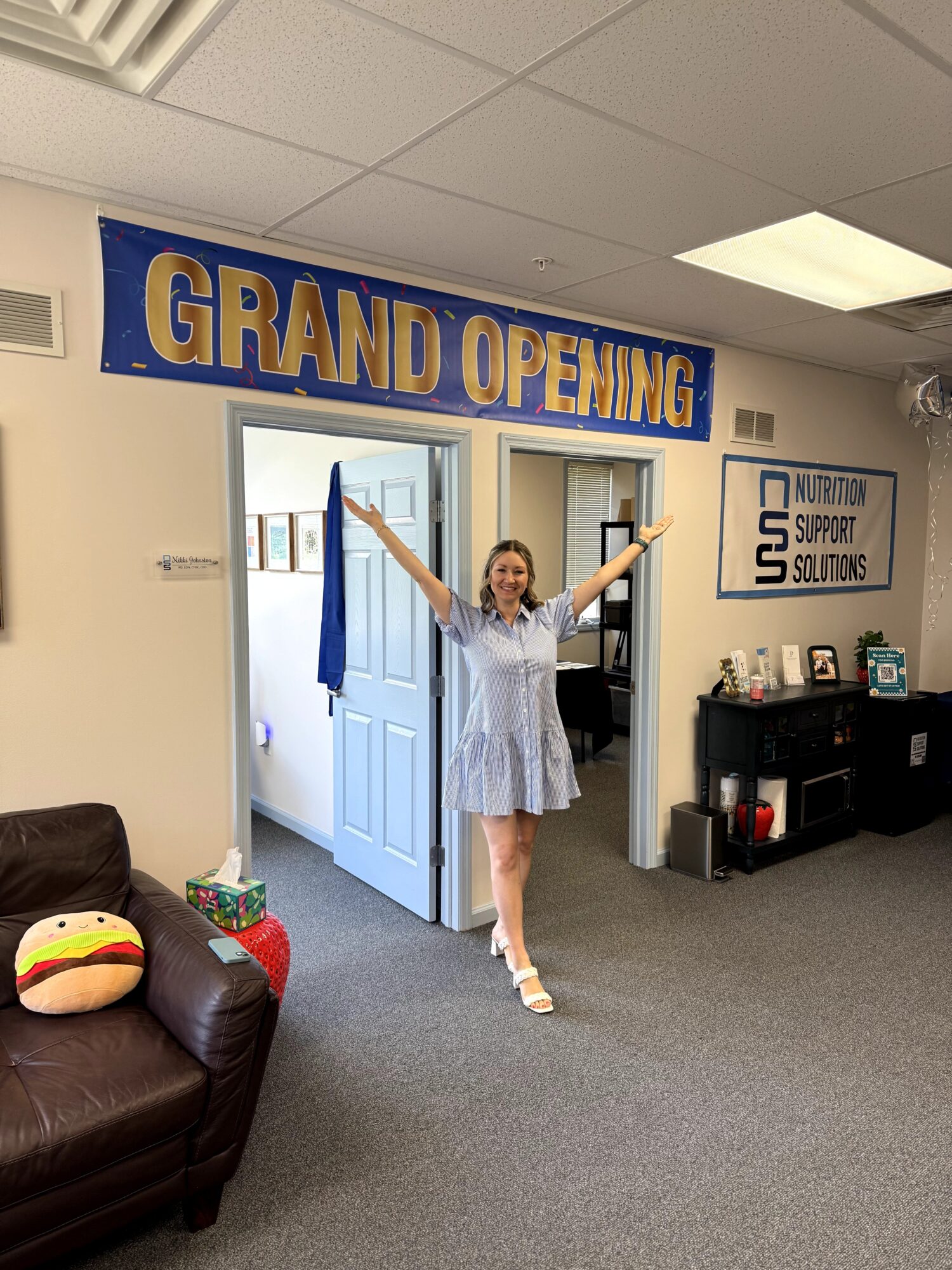 Young girl in a dress with arms raised standing in doorway under a grand opening banner, in an office space.