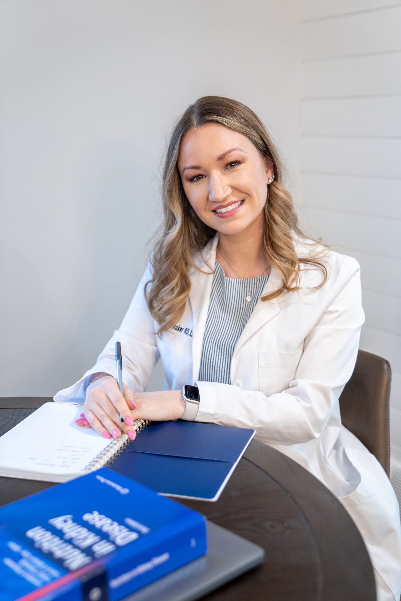 Smiling woman with wavy hair sitting at a table with notebooks and a pen, wearing a white blazer and gray top.