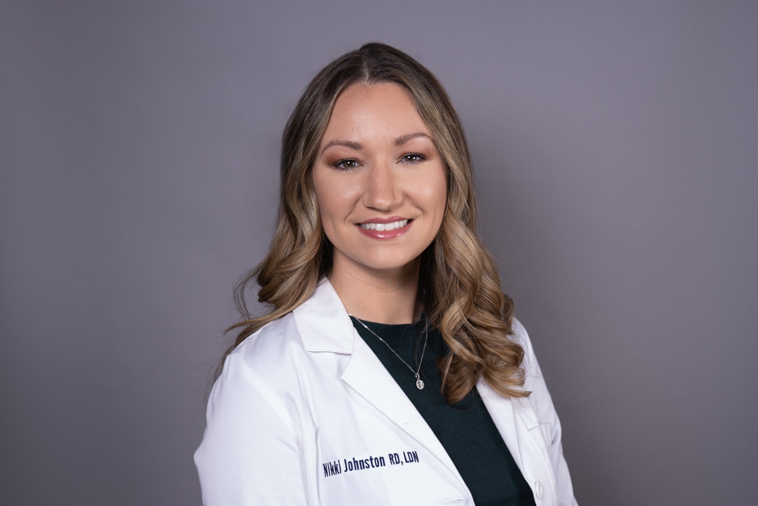 Young woman with wavy hair wearing a white lab coat and black top, smiling, against a gray background.
