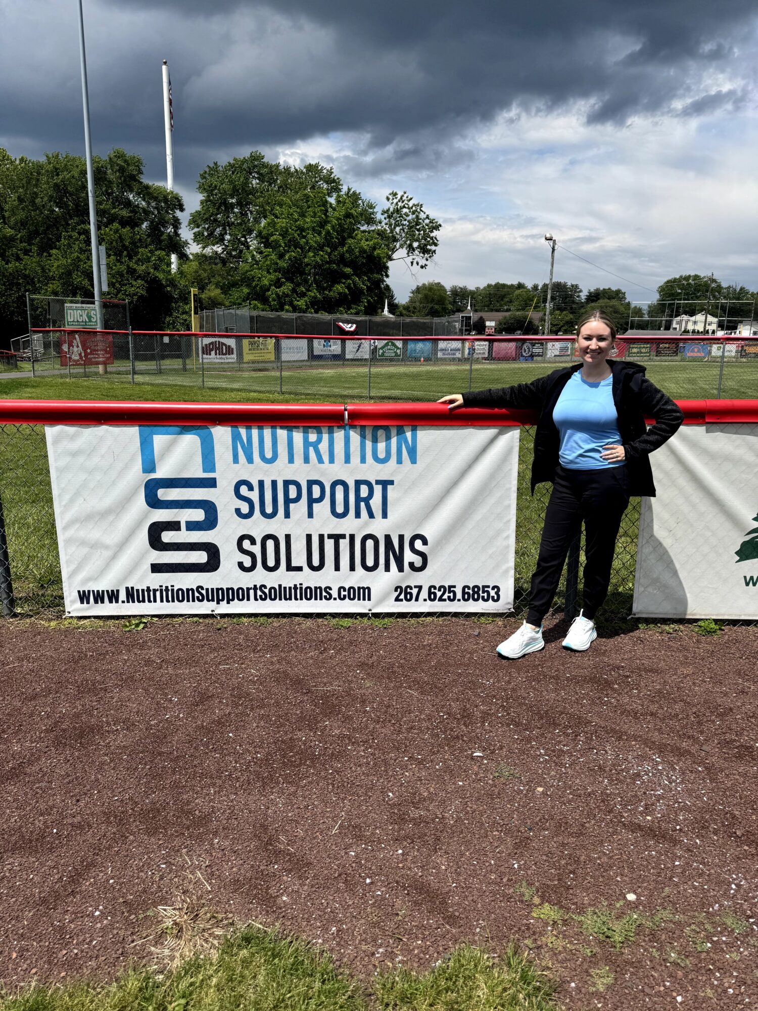 Person standing next to a banner on a sports field, cloudy sky, trees, and a fence in background.