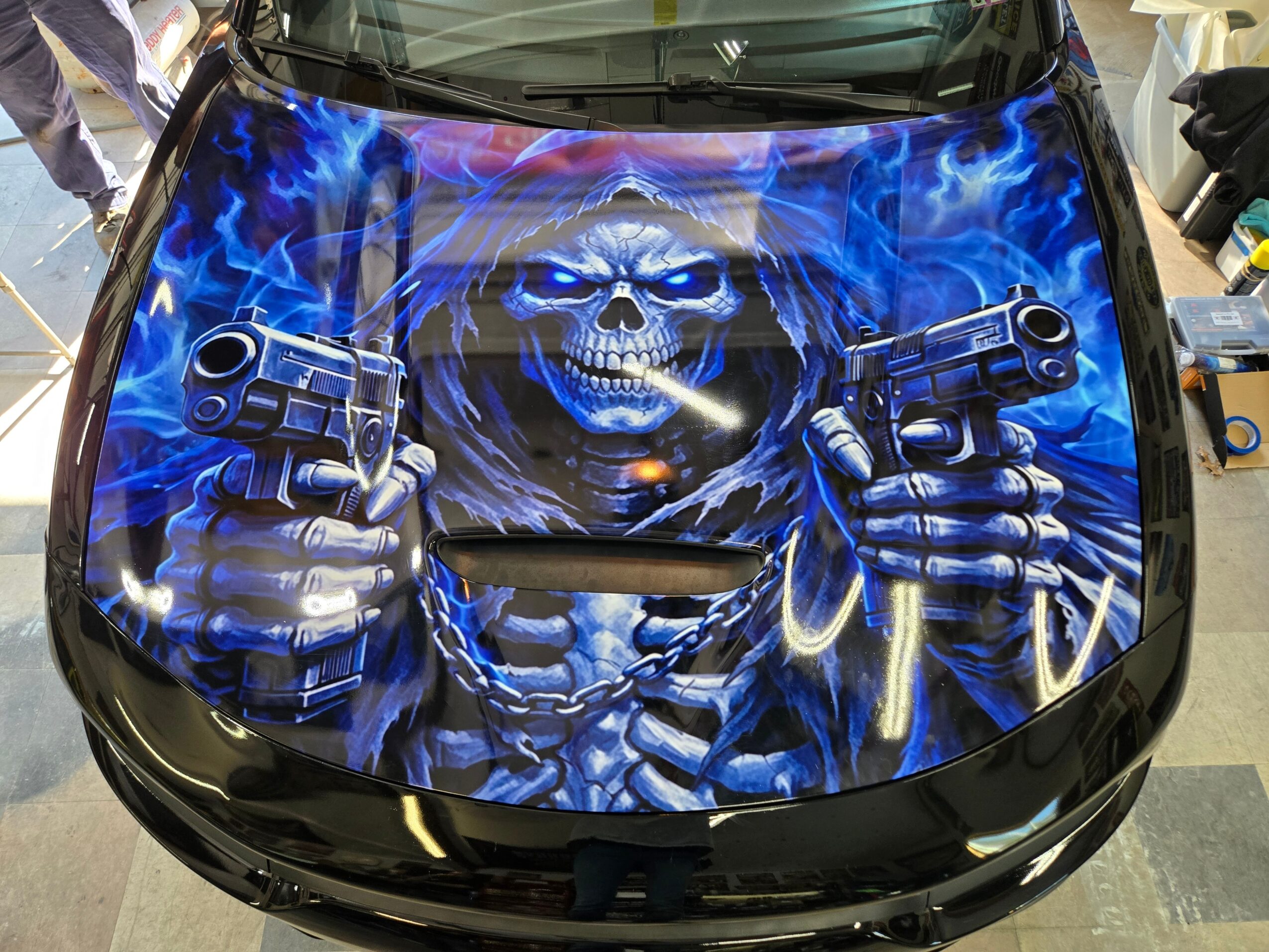 Car hood with a detailed airbrushed skull holding two guns, surrounded by blue flames and lightning effects.