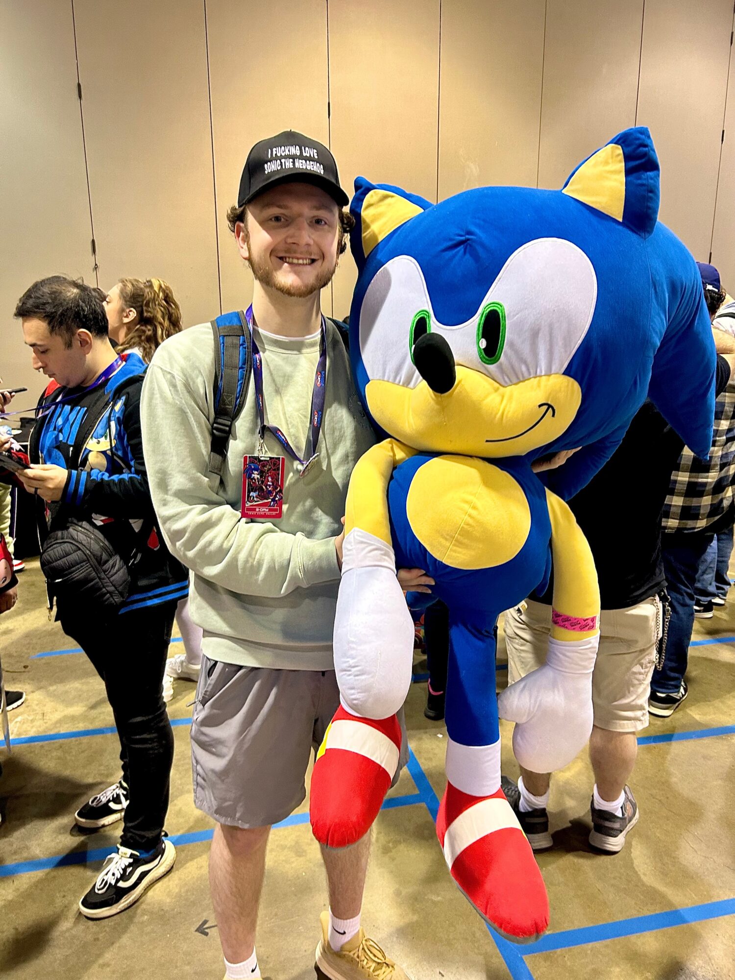 Person smiling next to a large Sonic the Hedgehog mascot at an indoor event, with other people in the background.