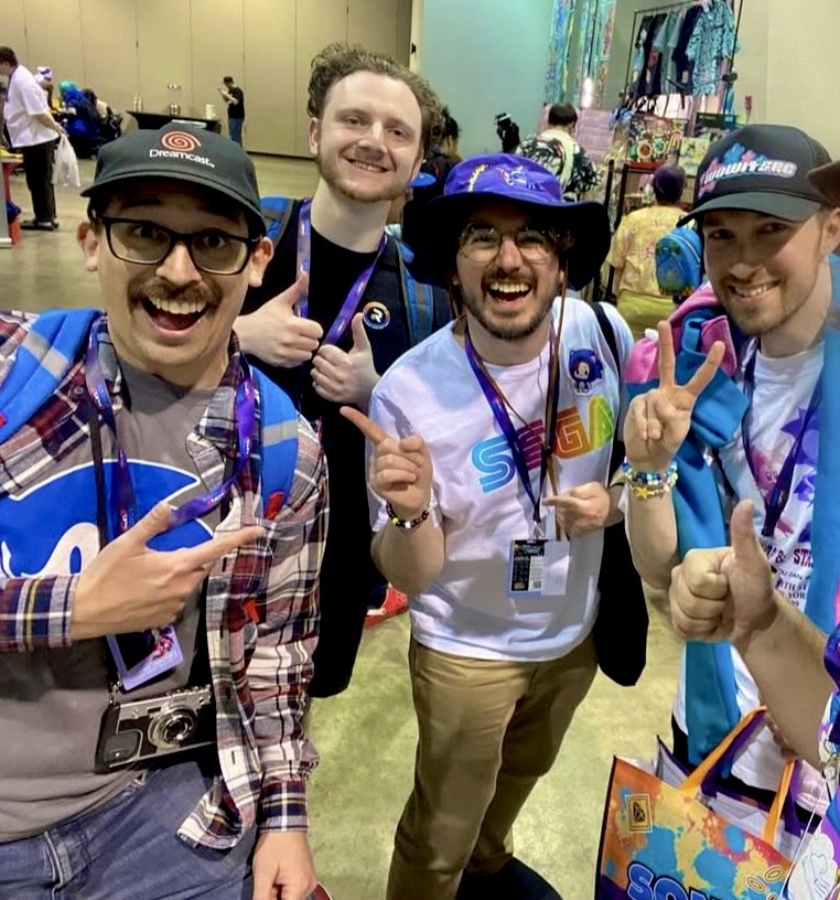 Four smiling men at an event, wearing hats and badges, making peace signs and pointing at the camera.
