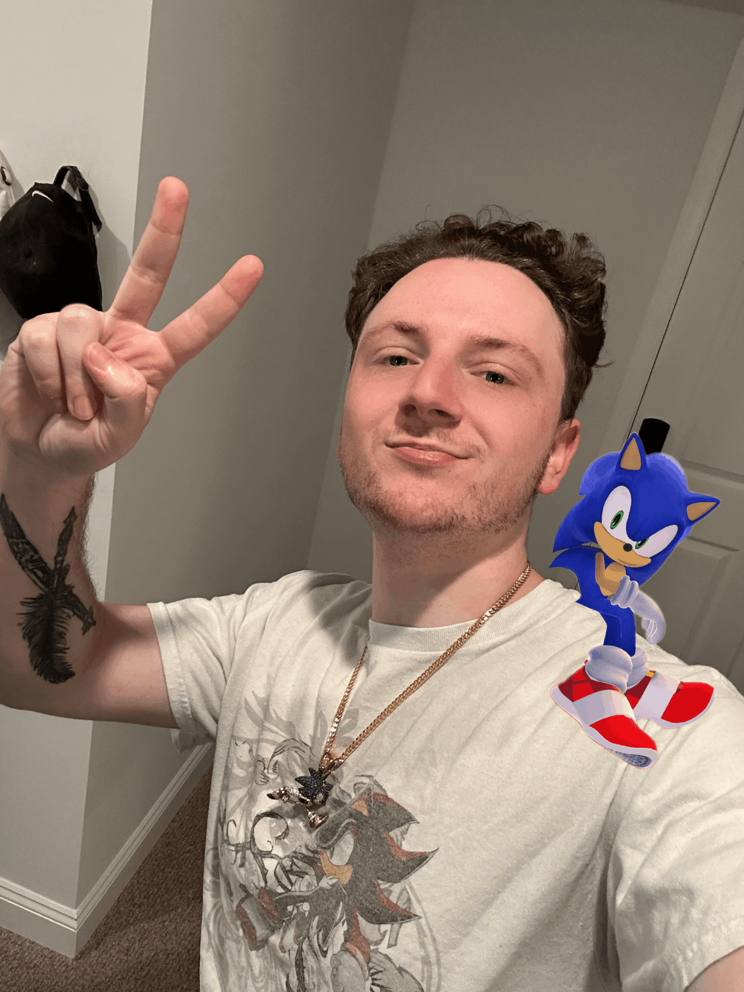 Young man with curly hair making a peace sign, wearing a white T-shirt with a graphic, and a Sonic the Hedgehog figure on shoulder.