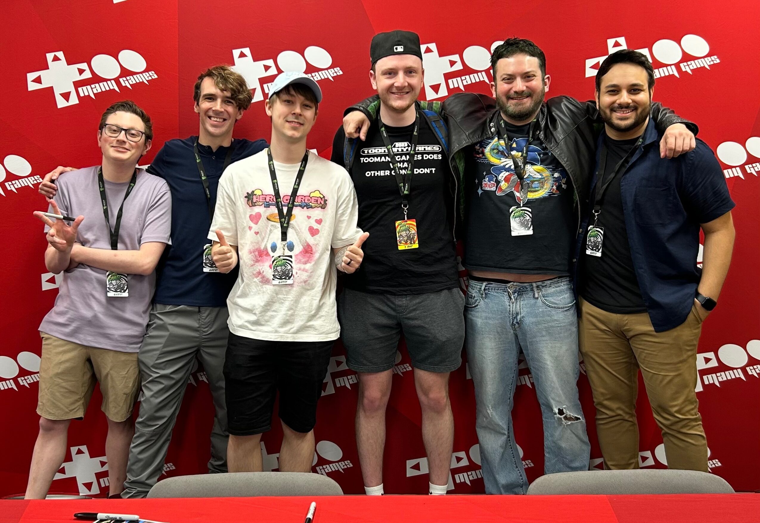 Group of six people standing together in front of a red backdrop with white logos, smiling and posing for the photo.