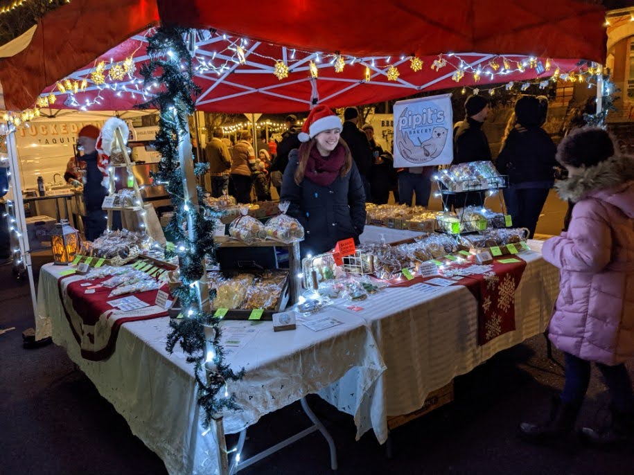 Christmas market stall with festive decorations, lights, and people browsing, including a woman in a Santa hat and a girl in a pink coat.