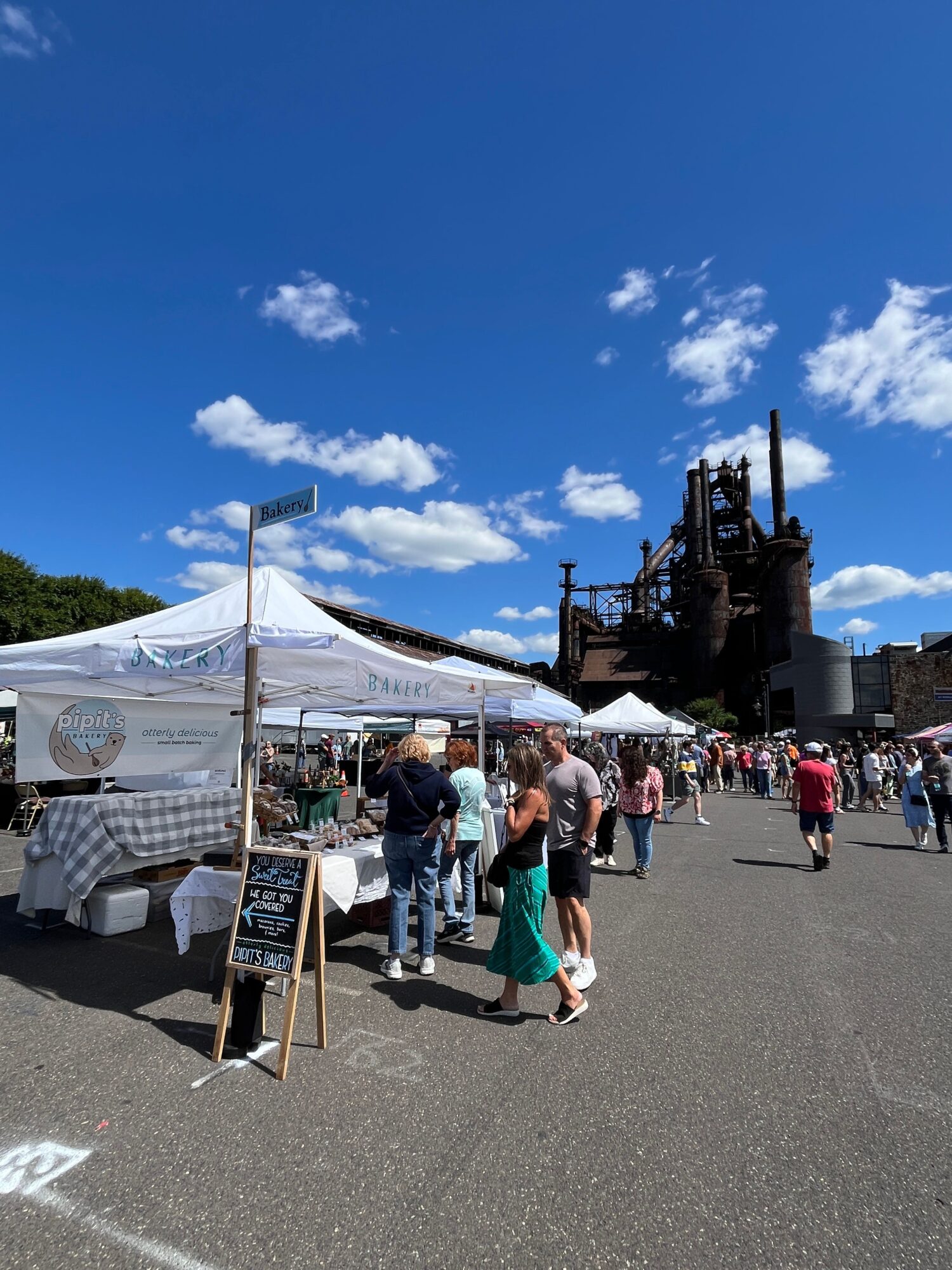 People gathered around a market stall with a white canopy, with a historic building and blue sky in the background.