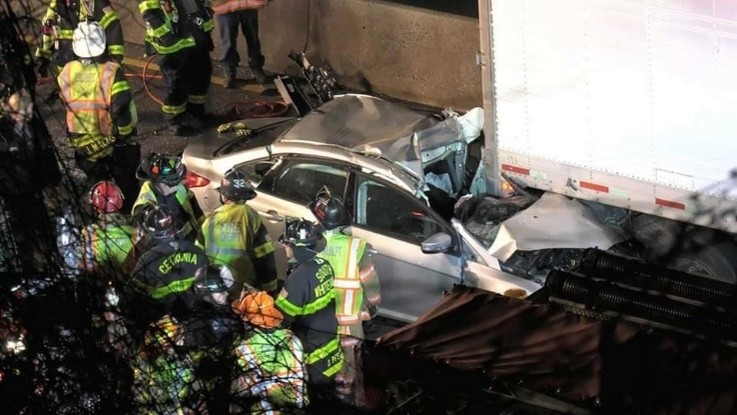 Car crashed into truck, emergency responders at the scene, some firefighters in yellow and black gear, in a dark area.