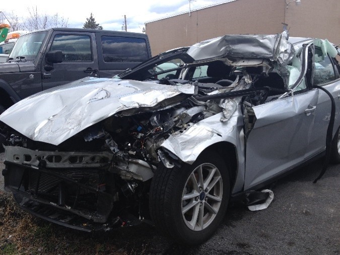 Damaged silver car with extensive front-end and roof damage, parked in lot with other vehicles, cloudy sky background.