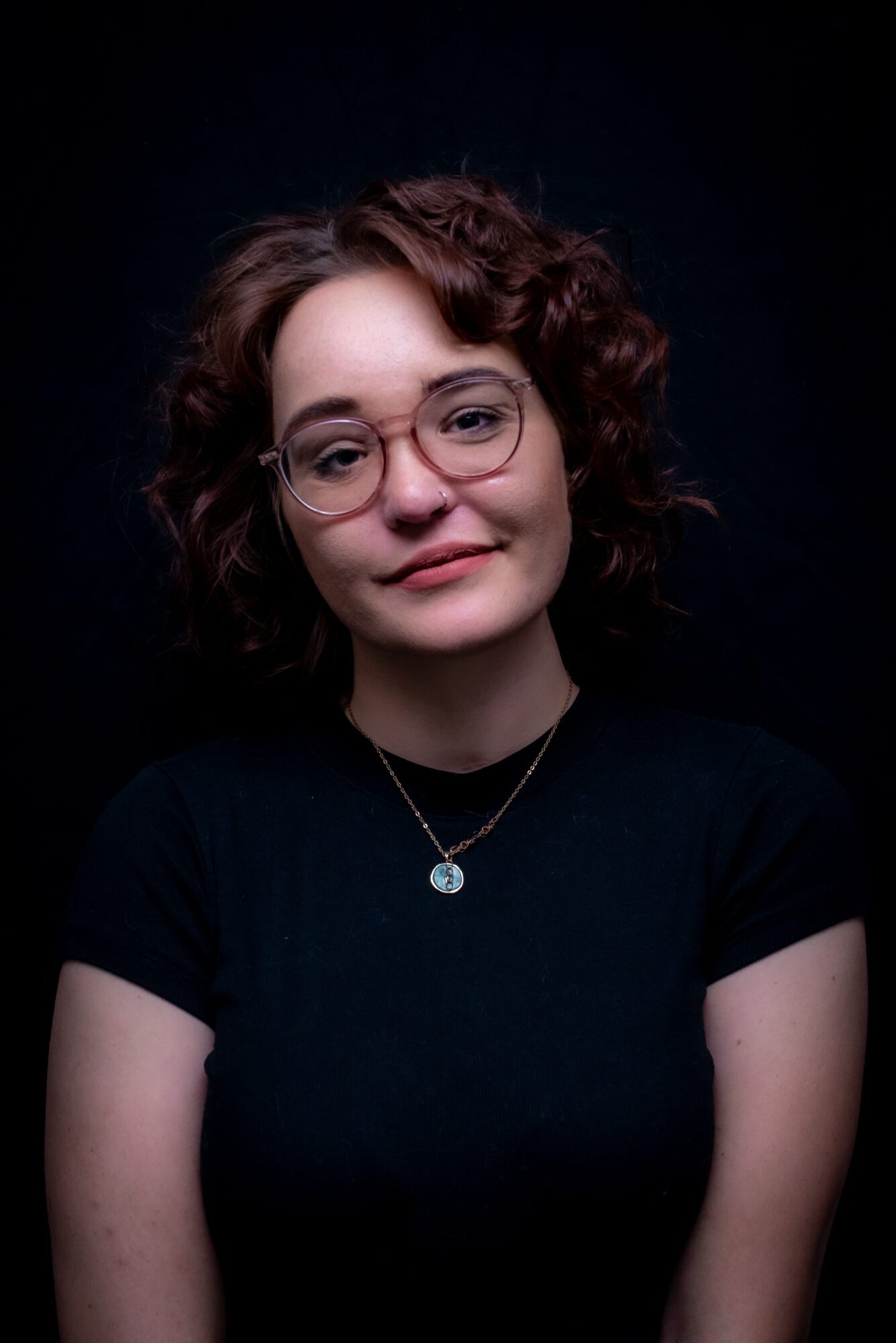 Young woman with curly hair and glasses, wearing a black shirt and necklace, against a dark background.