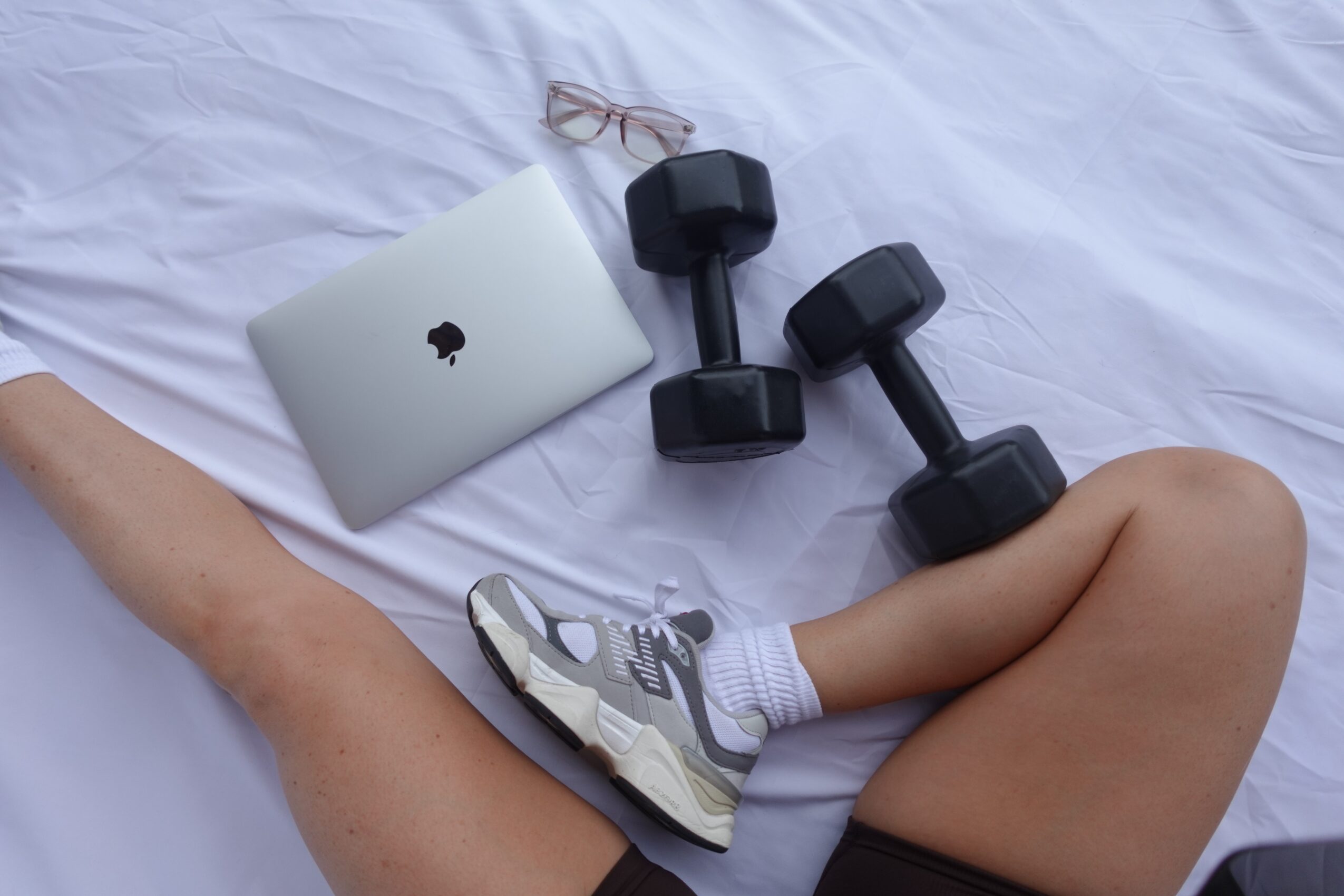 Person's legs with a sneaker, a laptop, a pair of dumbbells, and a pair of glasses on a white surface.