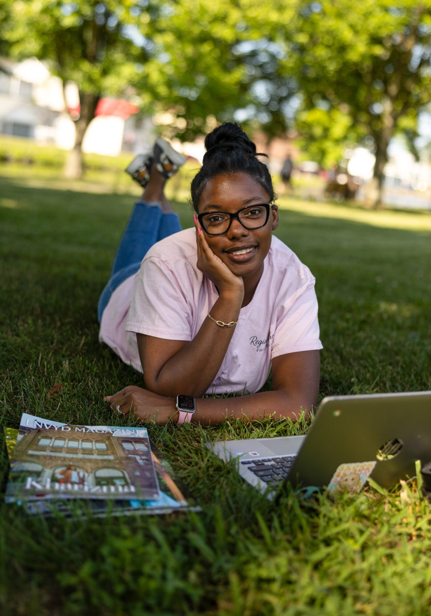 Young woman with glasses lying on grass outdoors, smiling, with a laptop and magazines nearby, trees in background.