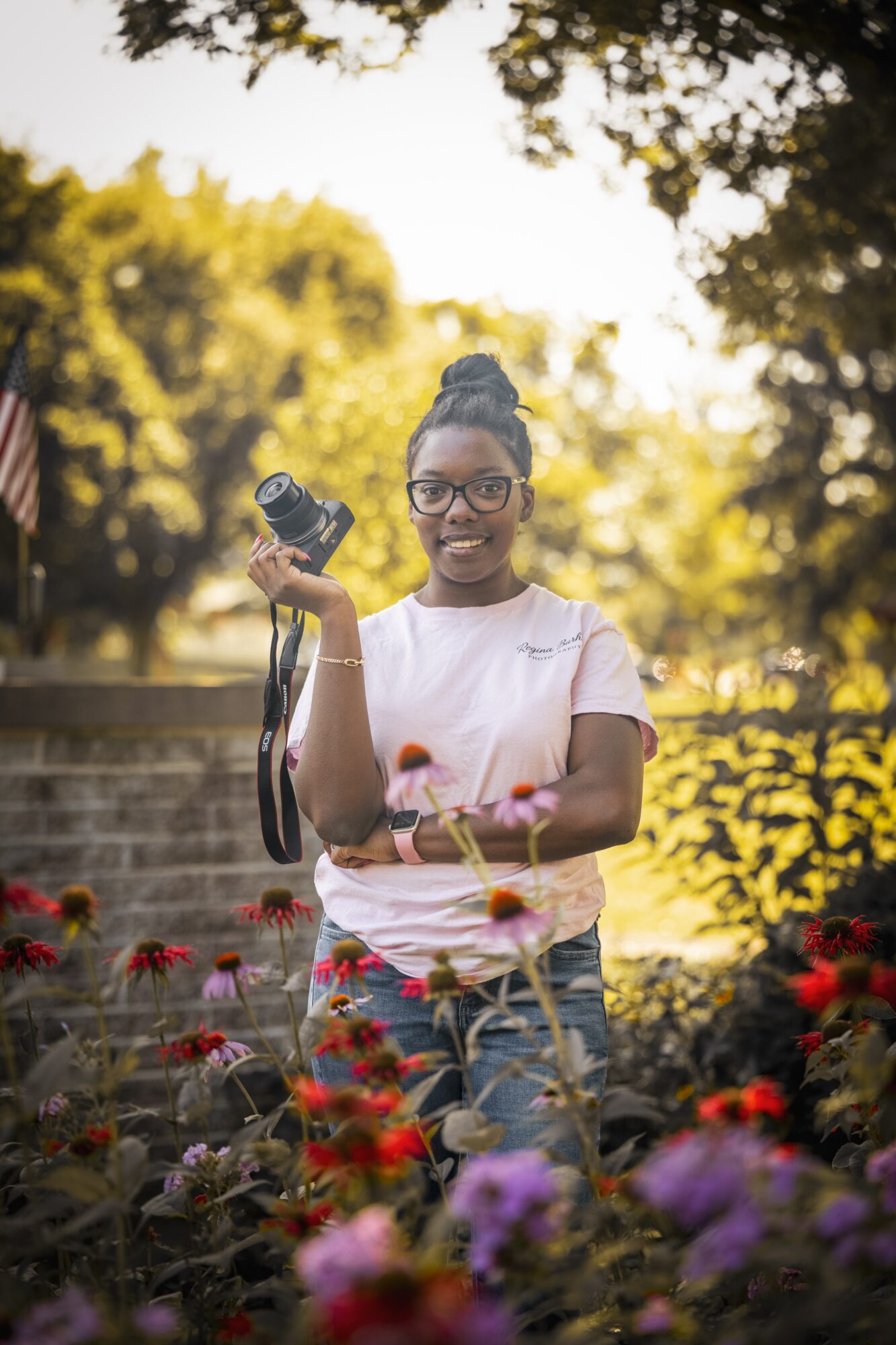 Young woman with glasses holding a camera outdoors in a garden with colorful flowers and trees.