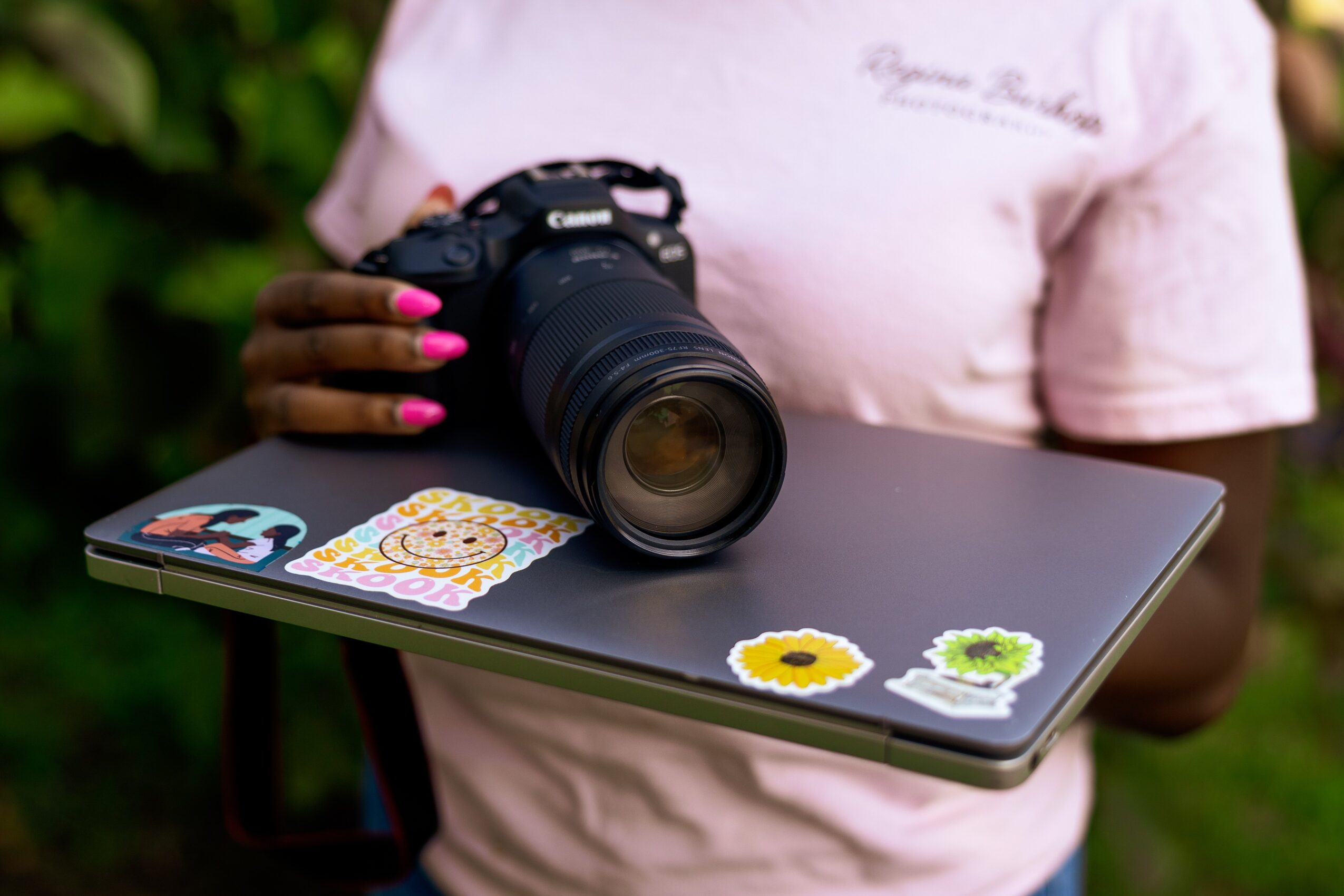 Person holding a camera and a closed laptop with stickers, outdoors with greenery in background.