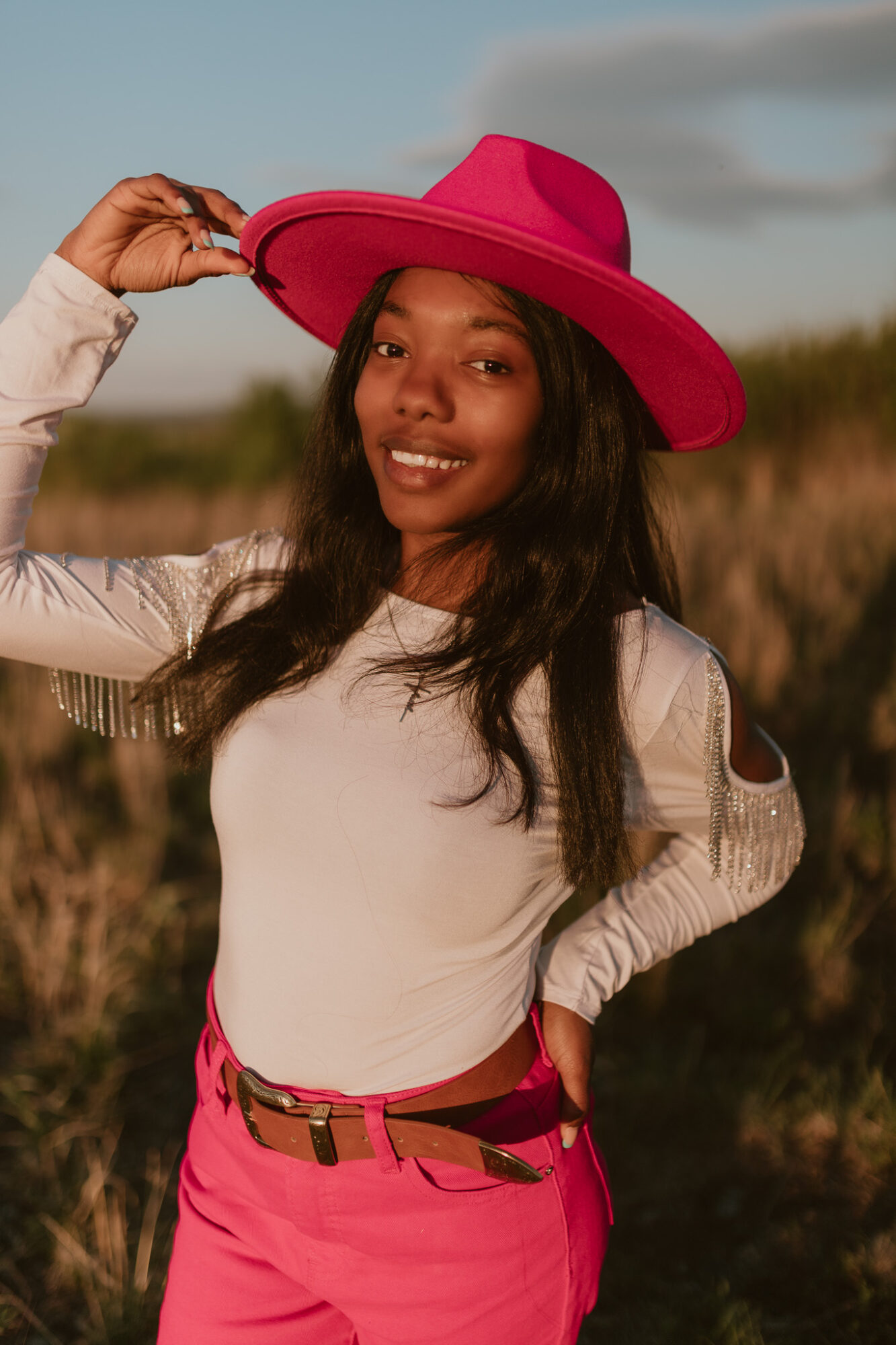 Young woman outdoors wearing a large pink hat, white top, and pink pants, smiling and holding her hat.