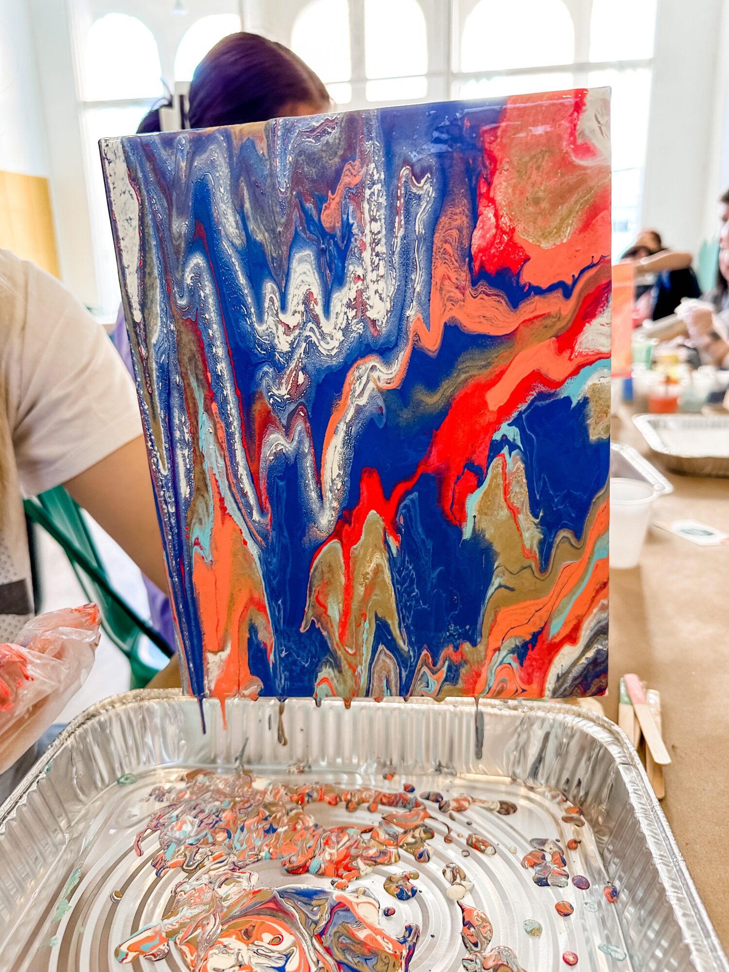 Person creating abstract painting with vibrant colors on canvas, with art supplies and people in background.