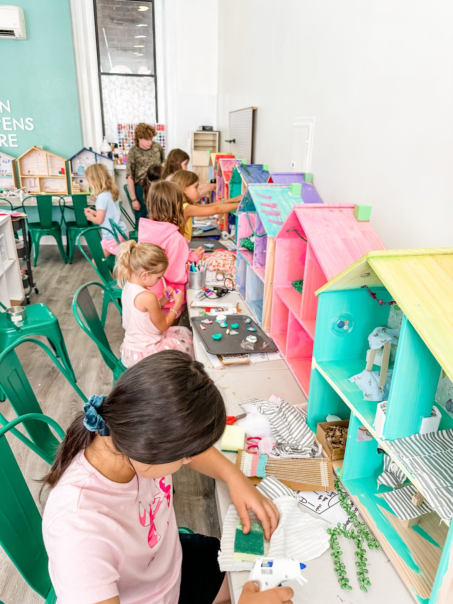Children at a craft table with colorful dollhouses and craft supplies, engaging in activities in a bright room.