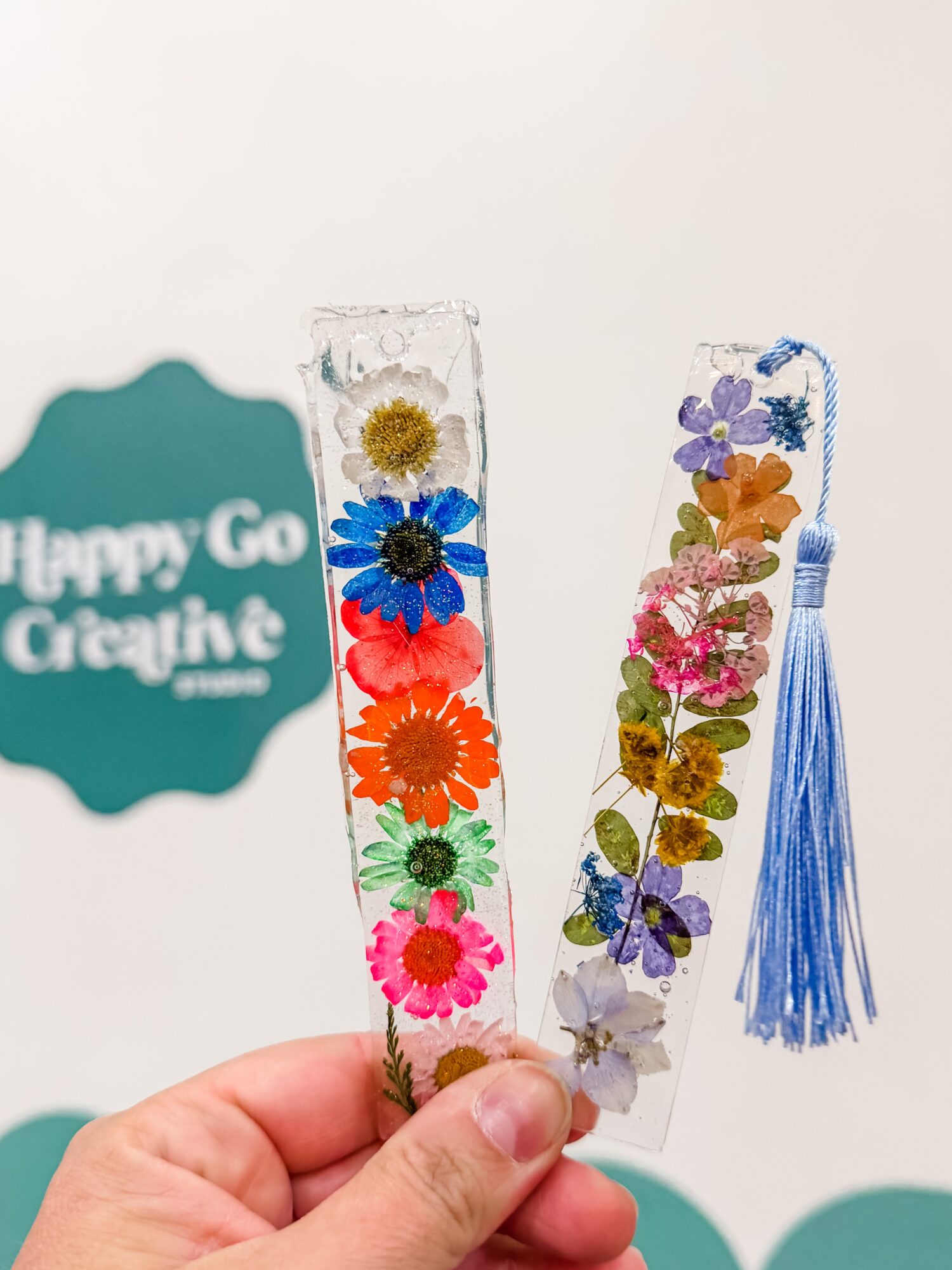 Two clear rectangular bookmarks with colorful pressed flowers and ribbons, held by a hand against a white background.