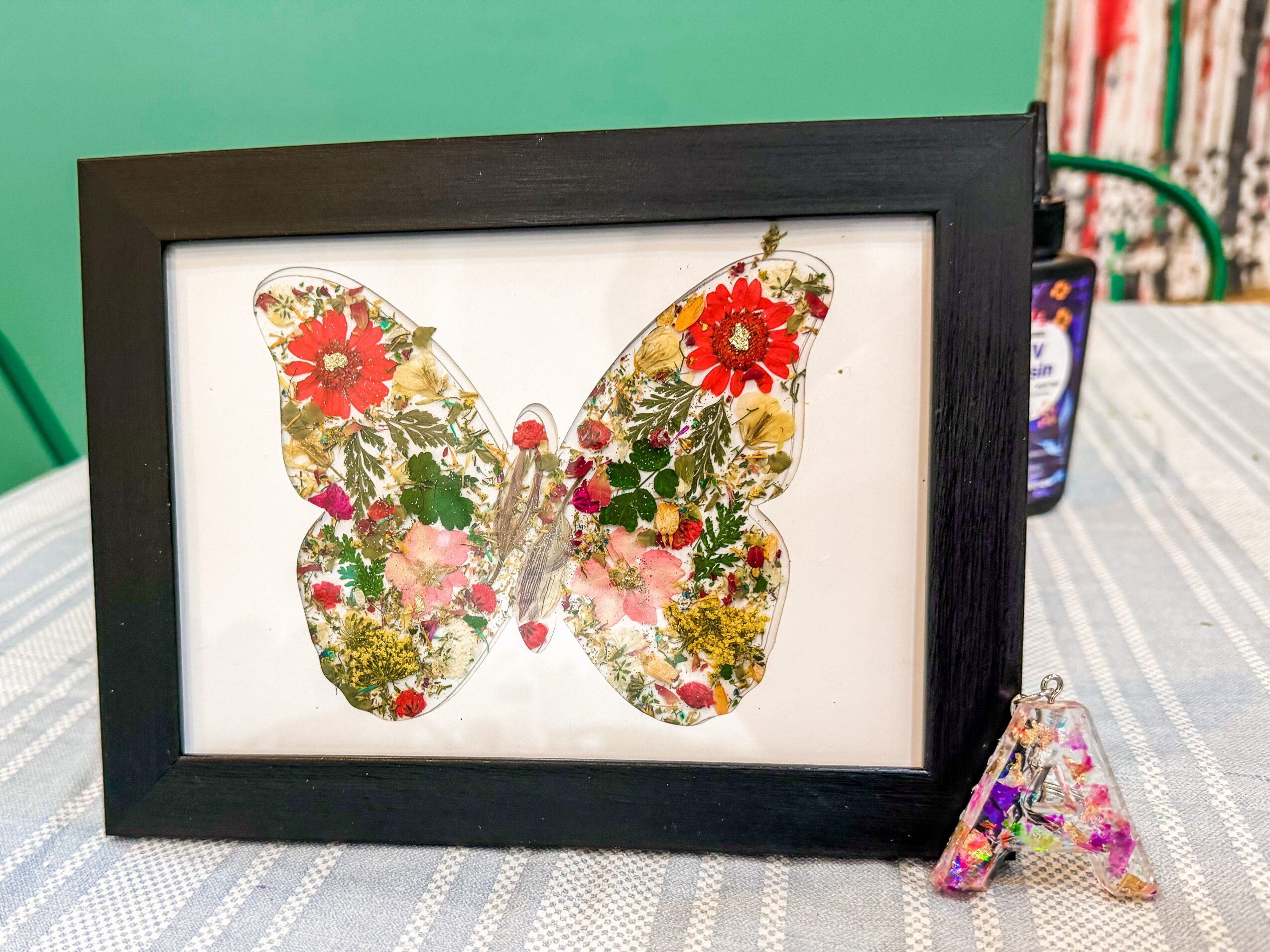 Framed butterfly artwork with colorful floral patterns, on a table with a letter A and a container in the background.