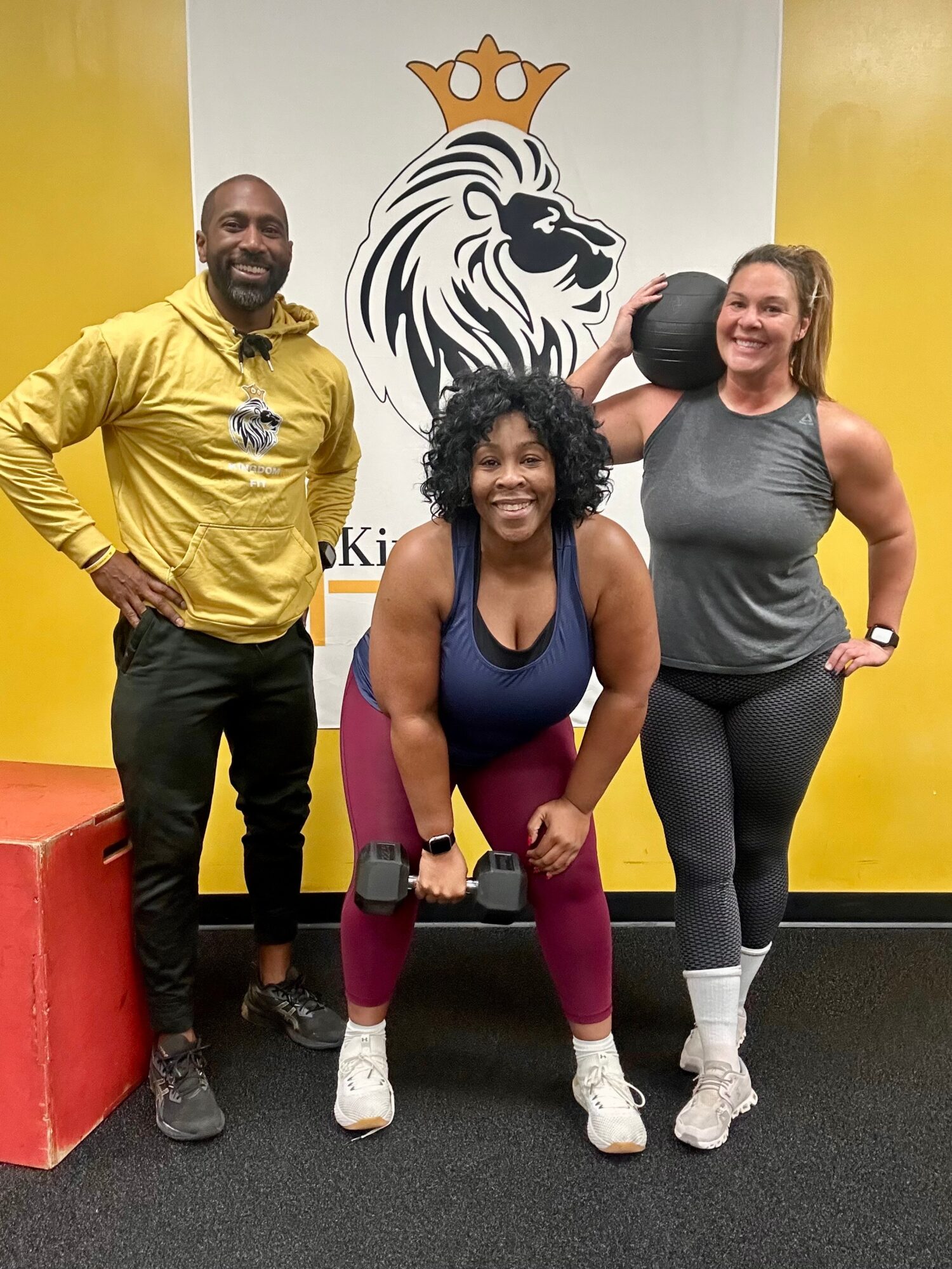 Three people in a gym, smiling, with a lion logo and crown on the wall behind them. One person is holding a kettlebell.
