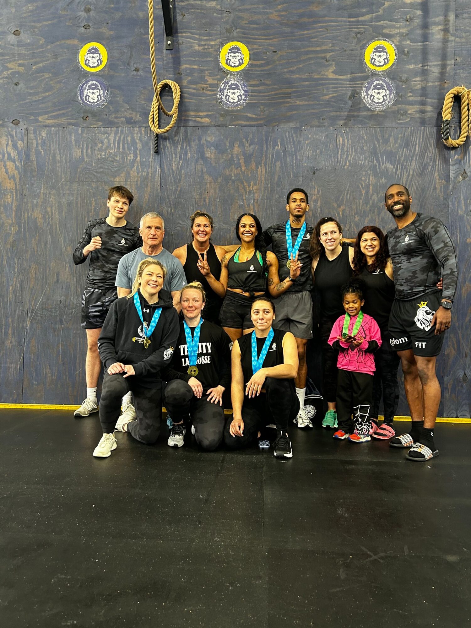Group of nine women and children posing indoors, some wearing medals, standing and sitting on a black floor with a dark wall background.